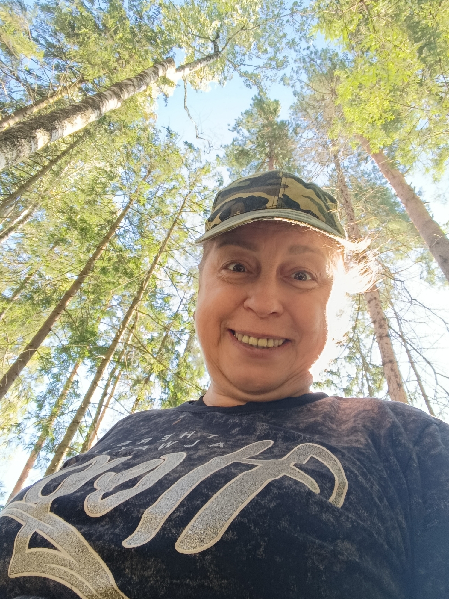 A woman is taking a selfie outdoors in a forest, looking up at the camera with a wide smile. She is wearing a camouflage baseball cap and a dark t-shirt with a graphic print. The background consists of tall trees with green foliage and a bright sky. The image appears to be taken in Zvorkovo, Russia.