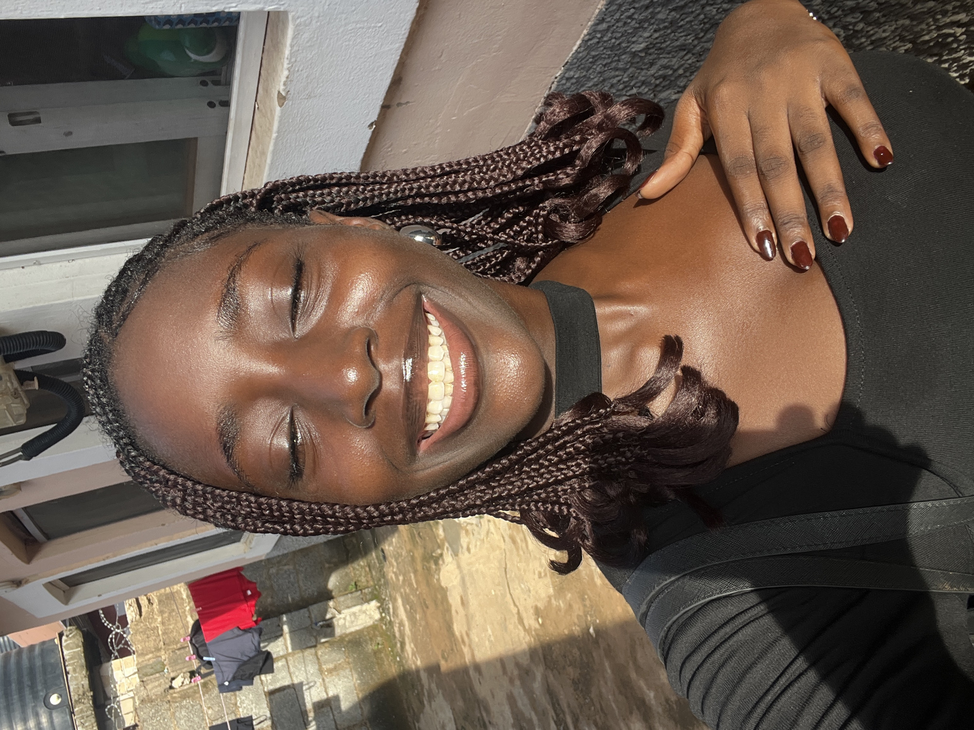 The image, rotated 90 degrees counter-clockwise, presents a close-up portrait of a young Black woman outdoors in Abuja, Nigeria, under bright sunlight. She has smooth, shiny dark skin and is wearing long, dark brown box braids with some curls at the ends. Her eyes are closed or squinting, and she is smiling broadly, revealing her white teeth.
Her left hand, with dark red nail polish, rests on her chest. She is dressed in a black top or dress with visible strap details and pleated fabric texture.
The background behind her head shows a light-colored building wall with a closed window. Below and to her side, an outdoor area with concrete or paved ground is visible. Further in the background, there appear to be clothes drying on a line, including a bright red item and some darker garments. The strong, direct sunlight suggests it is daytime, possibly midday or afternoon. The woman's bright smile conveys an emotion of happiness or joy. No visible text is present in the image.