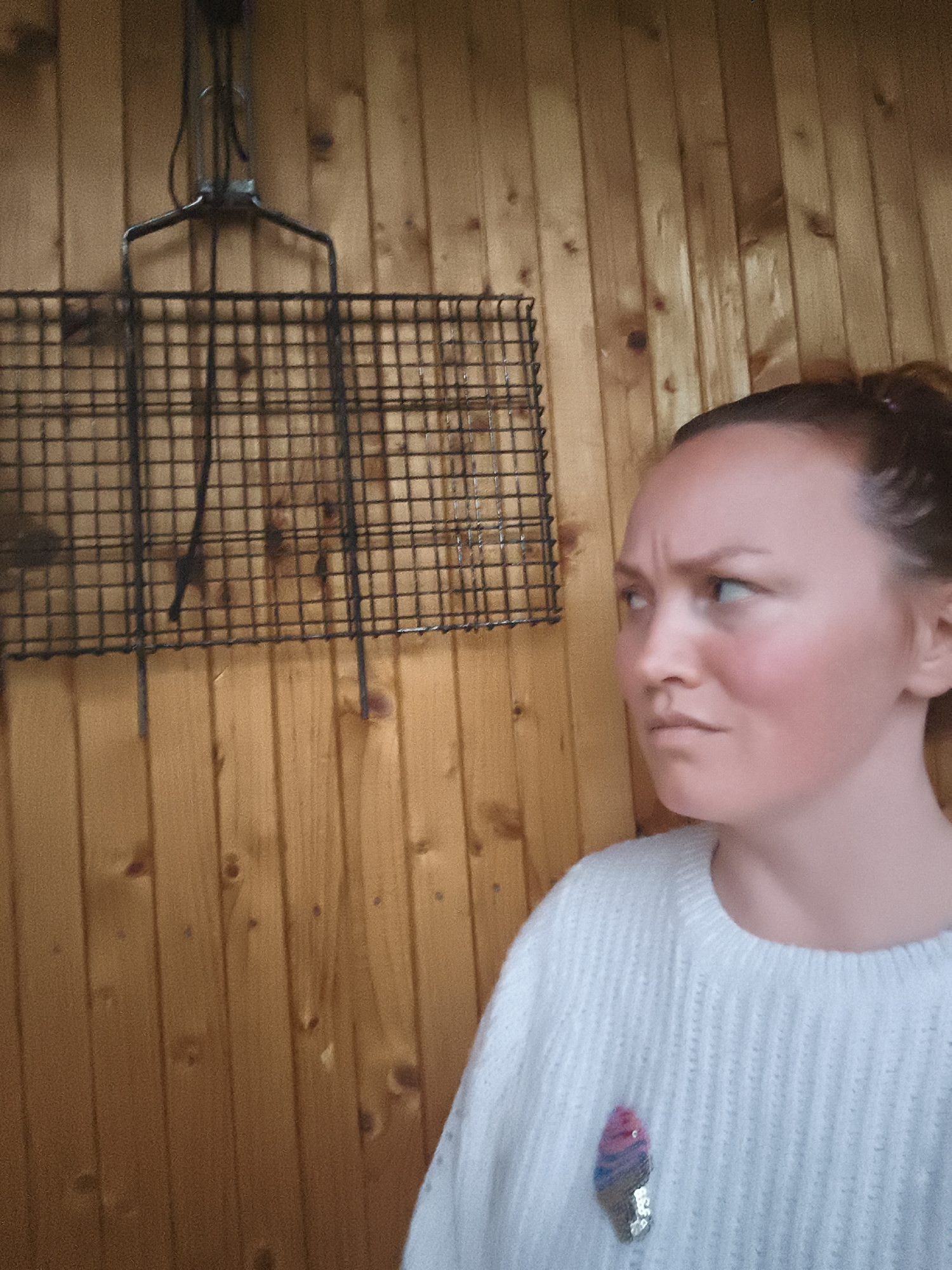 In an unknown location within the city of Unknown, Unknown, a frontal profile shot captures a light-skinned female individual from the chest up, positioned against a vertical wooden plank wall. The individual, estimated to be a young to middle-aged adult, has dark hair pulled back. Her face displays a furrowed brow, indicating a stern, displeased, or concerned expression, with her gaze directed intently towards the left of the frame. Redness is visible across her cheeks and forehead. She is wearing a light-colored, ribbed knitted sweater adorned on the left side with a sequined pin shaped like an ice cream cone, featuring pink, blue, and silver sections. To the left of the individual, a dark, rectangular metal grid structure, resembling a grilling rack, hangs against the wooden wall, suspended by a metal fixture at its top. The wooden wall itself consists of vertically aligned planks with visible grain and knot patterns.