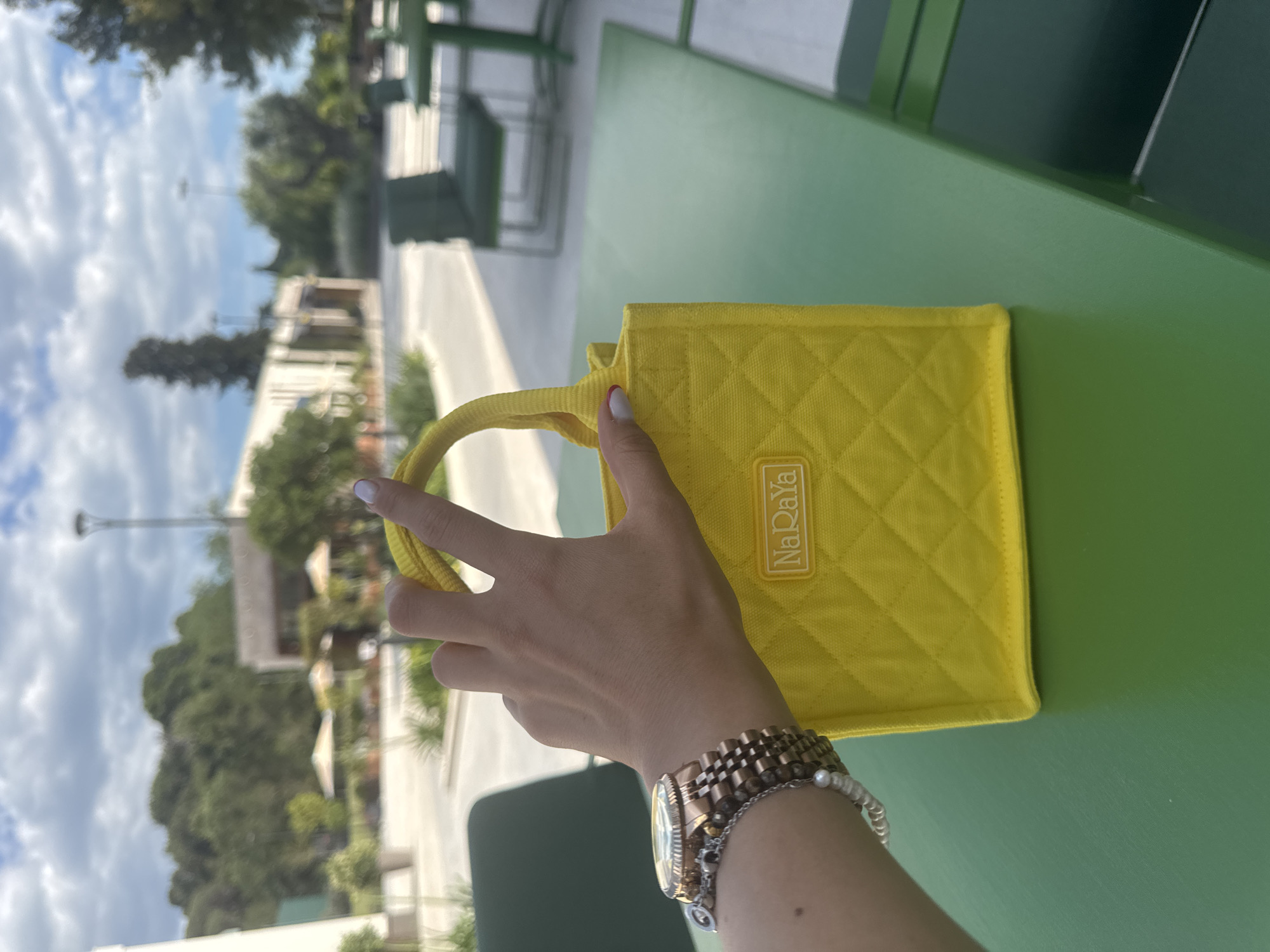 The image shows a person holding a yellow quilted NaRaYa handbag outdoors in Tivat, Montenegro. The background reveals a partially obscured cityscape with light-colored buildings, trees, and a cloudy sky. A green metal table is in the foreground, extending behind the bag. The person is wearing a watch and bracelets on their left wrist, partially visible. The handbag has a diamond quilted pattern and the "NaRaYa" logo.