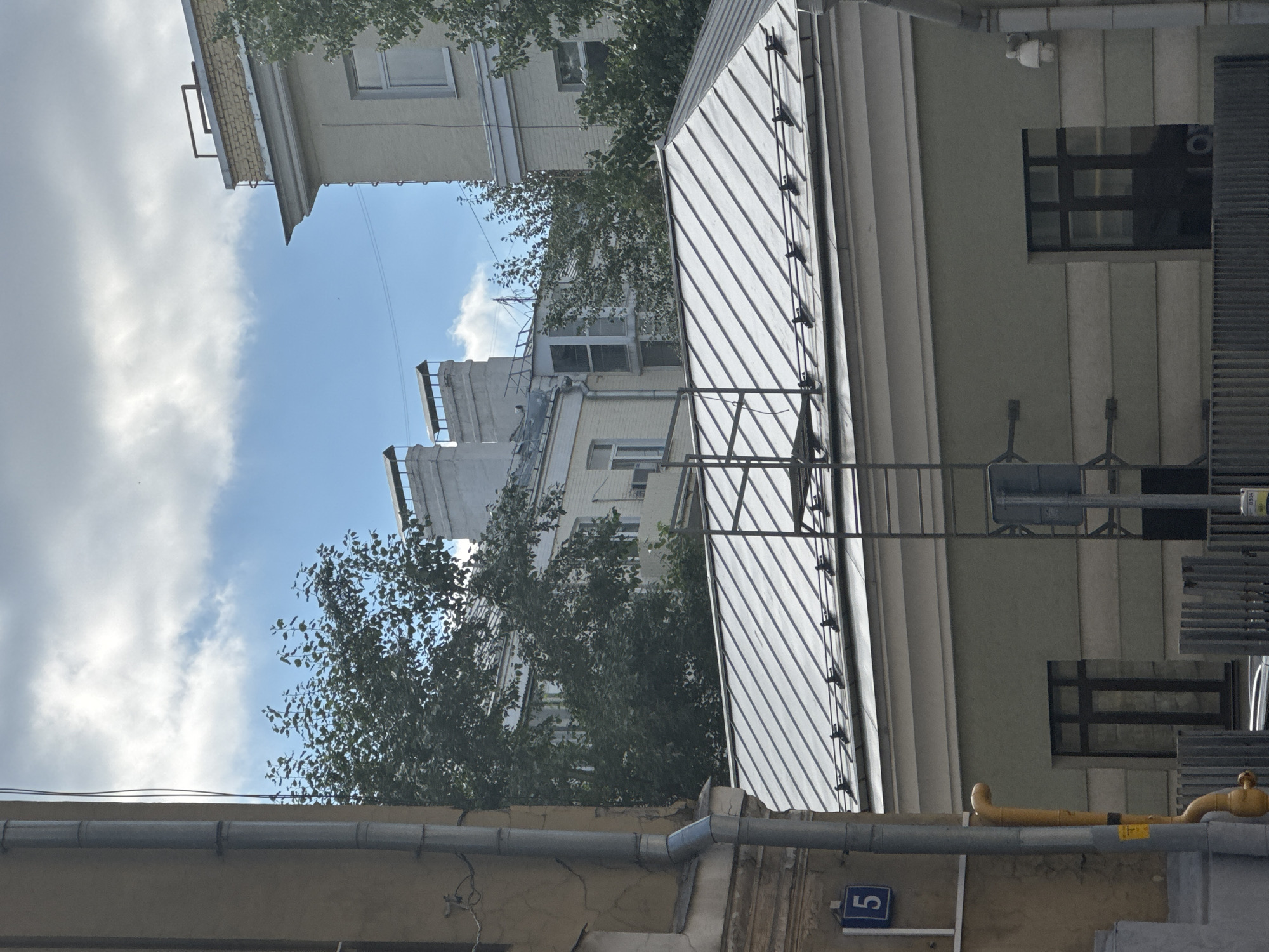 The image shows a view of buildings in Moscow, Russia. In the foreground, a light-colored building with a gray metal roof is visible. The roof has a series of parallel seams and is supported by a structure with metal railings. Below the roof, the building has a light green facade with horizontal banding and a dark-framed window. To the left of this building, a tall, gray downspout runs vertically along a cream-colored building, with a blue sign showing the number "5" attached to it. A yellow pipe is also visible attached to the cream-colored building.
In the middle ground, behind the foreground building, several other buildings are visible, some with light yellow facades and white trim. These buildings have chimneys with distinctive rectangular tops. Trees with dark green foliage are interspersed among the buildings, obscuring some of the architectural details.
In the background, the sky is blue with scattered white clouds. The overall scene depicts an urban environment with a mix of architectural styles. The lighting suggests it is daytime, with the sun casting some shadows. There are no people or animals visible in the image.