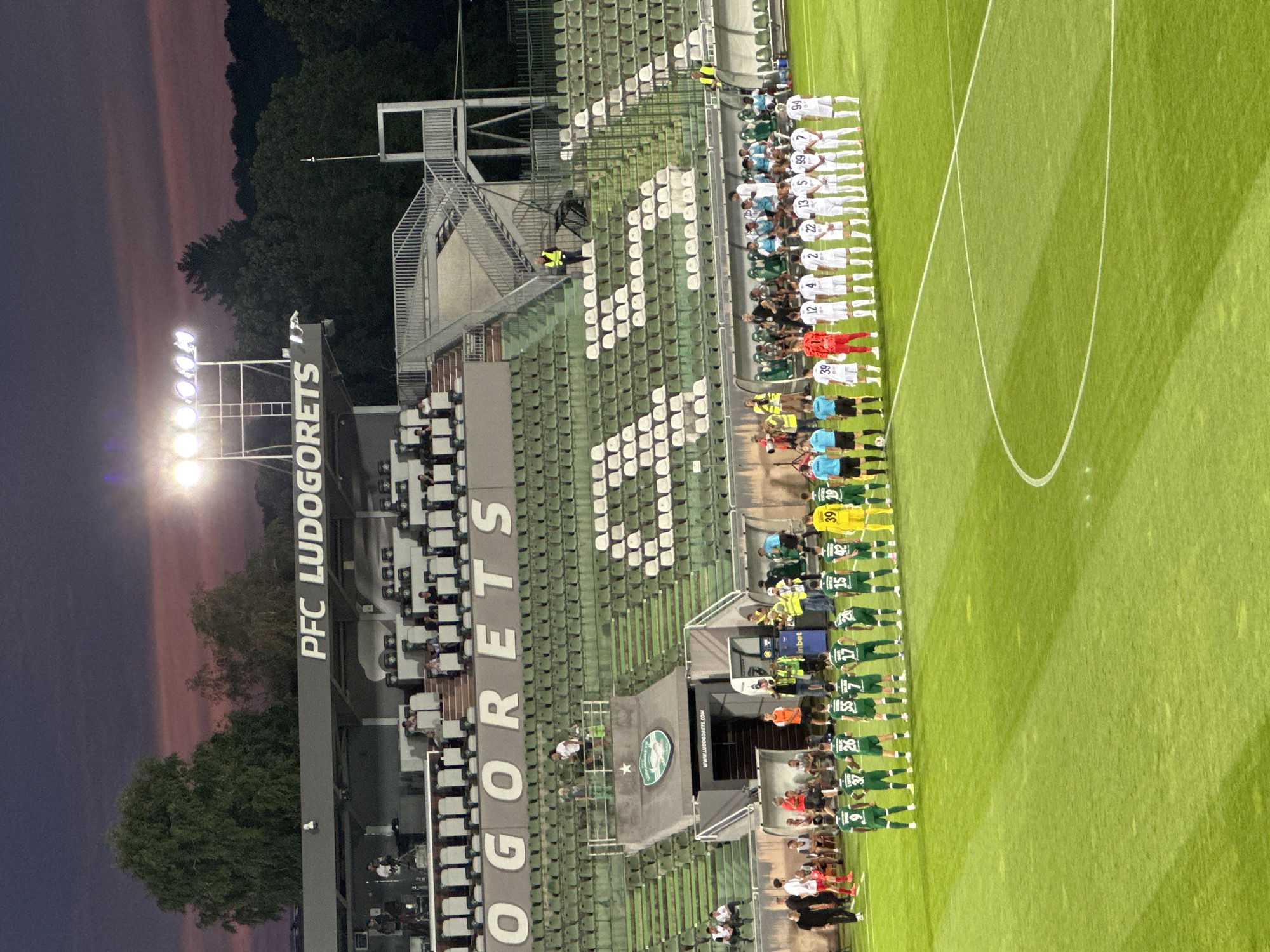 A football match is about to begin at the Huvepharma Arena, located in Razgrad, Bulgaria. The stadium is illuminated by floodlights, with a deep purple and pink sunset visible in the sky. Two teams of players are lined up on the green football pitch, facing each other. One team is wearing green kits, and the other is wearing white kits. Spectators are seated in the tiered stands behind them. The stadium features prominent signage displaying "PFC LUDOGORETS."