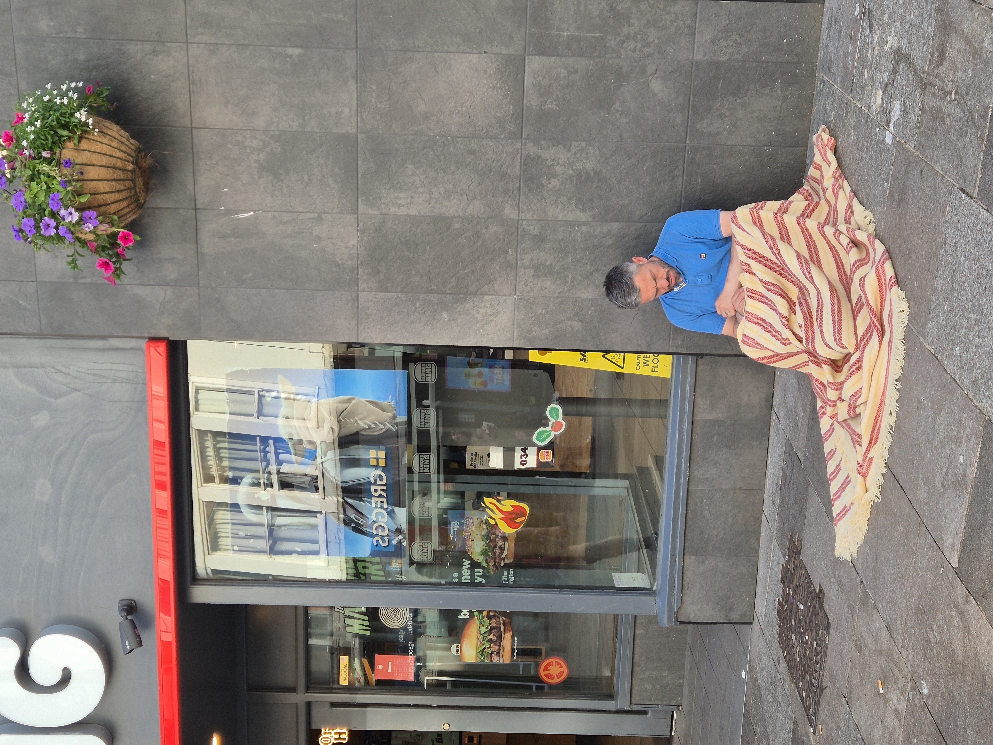 The image shows a man sitting on the sidewalk in front of a building in Cardiff, United Kingdom. He is wearing a blue polo shirt and has a blanket wrapped around him. A hanging flower basket is attached to the building to the left of the man. A Burger King restaurant is to the left of the man and flower basket. The building wall is grey. The sidewalk is made of grey stone slabs.