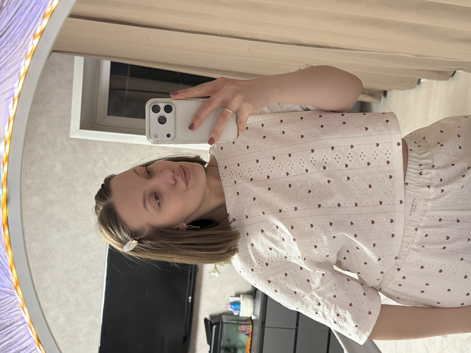 A young woman is depicted taking a mirror selfie in an indoor setting in Reshetnikovo, Russia. The image is oriented sideways, but depicts her standing upright, holding a light-colored smartphone with three camera lenses in her right hand, angled towards the mirror. She has light brown, shoulder-length hair, with a white and gold barrette clipped on the right side, and a neutral expression as she looks at the phone. She wears a matching set consisting of a white, short-sleeved top and shorts, both featuring a pattern of small red hearts and eyelet embroidery. A simple gold band ring is on her right ring finger, and her nails are painted a light plum color.
The large, oval mirror she is using has an illuminated, patterned frame on its left side. The reflection in the mirror reveals a room with light, textured wallpaper. In the background, light brown curtains are drawn over a window. Below the woman's head, a dark-screened television is visible, beneath which sits a dark cabinet holding a small container (possibly an aquarium) and a white artificial flower. The ambient lighting and the illuminated mirror frame suggest it is likely evening or night. A white label on her shorts is visible in the reflection, showing the reversed text "PRIMWEЯS," which translates to "PRIMWERS."