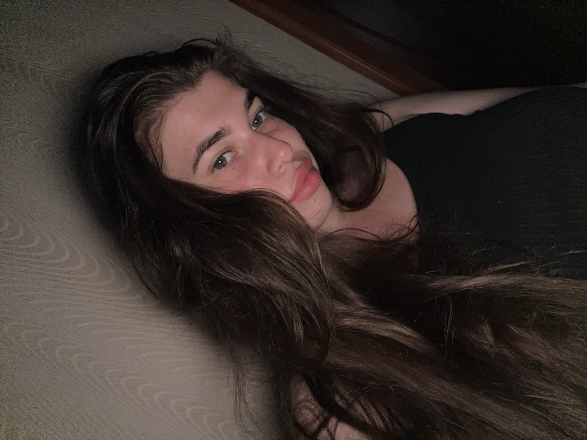 This is a close-up, dimly lit selfie of a young woman. She has long, dark brown hair that is slightly messy and falls around her shoulders and face. Her eyes are open and looking towards the camera, and she has a soft, pouty expression with slightly parted lips. Her skin appears fair with a pinkish hue on her cheeks. She is wearing a dark, ribbed tank top. The background consists of a textured wall with a wood-grain pattern, and a dark area to her right, possibly a doorway or shadow. There are no discernible urban elements or indicators of the city of Мещёры, Russia. The overall lighting is low, creating a moody atmosphere.