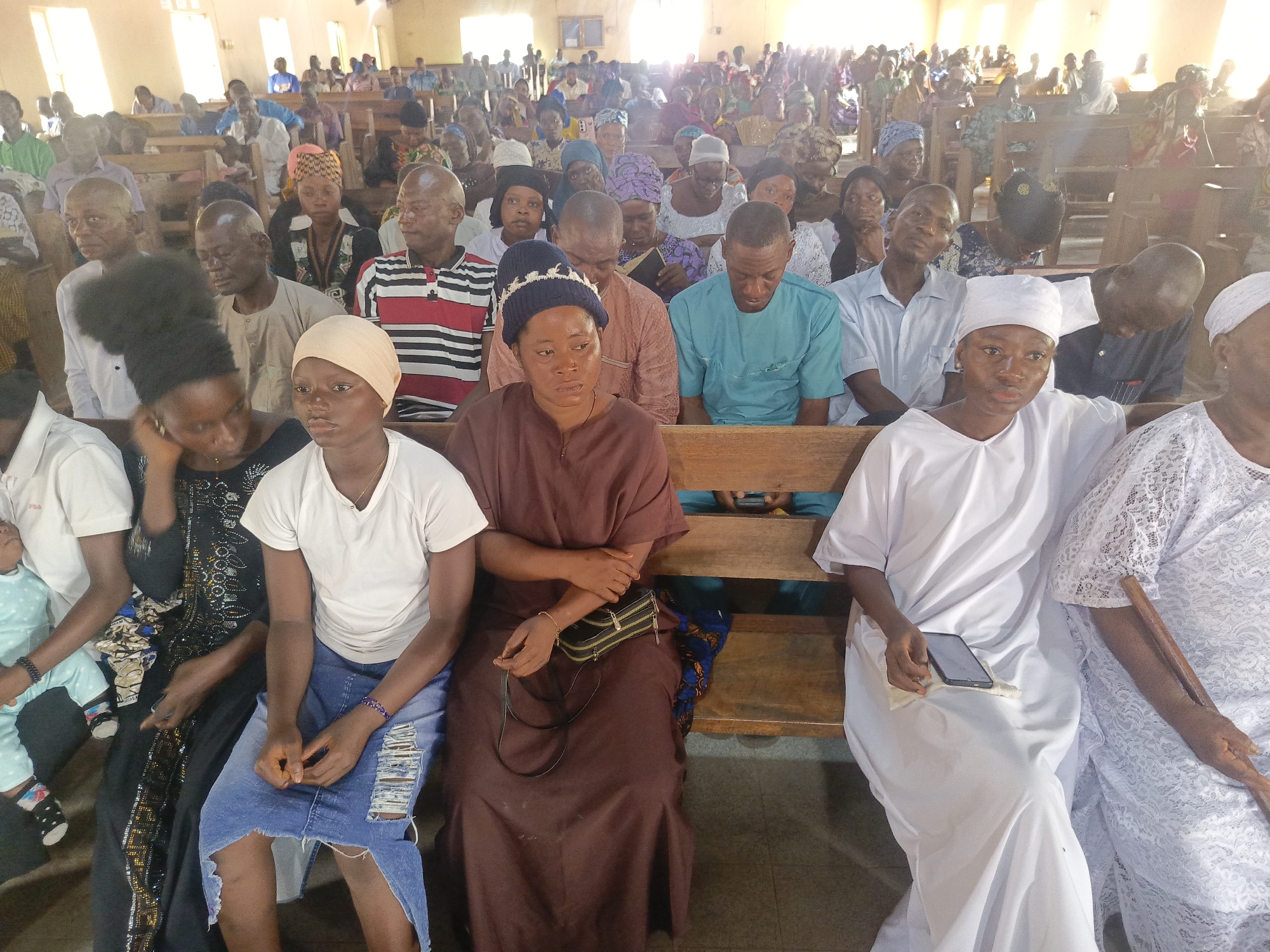 This image depicts a large gathering of people, mostly adults, seated on wooden pews inside what appears to be a church or community hall in Akwanga, Nigeria. The hall is brightly illuminated by strong natural light streaming in from windows on the upper right side, suggesting it is daytime.
In the foreground, several individuals are clearly visible. On the far left, a woman wearing a black dress with embellishments is seated next to a baby dressed in a light blue, polka-dotted outfit. Beside them, a young person with a light brown headwrap, wearing a white t-shirt and a distressed denim skirt, looks forward. The central figure in the front row is a woman wearing a dark blue knit cap over a headwrap, a long brown dress, and carrying a small black shoulder bag. To her right, another individual in a white headwrap and a white long-sleeved garment holds a smartphone and a white cloth. On the far right, a woman in a white lace dress is visible.
The mid-ground and background are densely populated with many more attendees, seated in rows. Their clothing is diverse, including traditional African patterns, various head coverings for women, and casual shirts. Many of the men have closely shaved heads. Most people appear to be looking towards the front or down, with generally calm and attentive expressions. The overall scene suggests a religious service, community meeting, or similar formal gathering. No discernible text is visible anywhere in the image.
