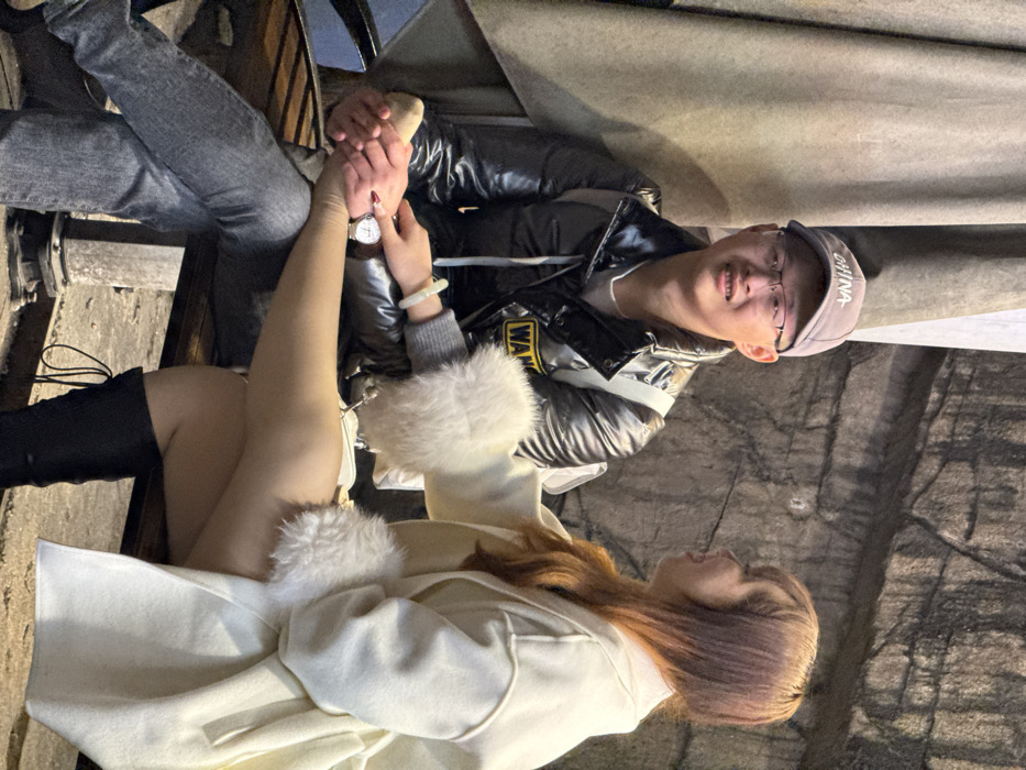 A couple is sitting closely together. The man is wearing a shiny puffer jacket, jeans, and a baseball cap, and is smiling at the camera. The woman, wearing a cream-colored coat with fur trim and tall black boots, is looking up at the man, holding his hand. They are in an outdoor setting, possibly in a cave or a tunnel. The lighting is dim.