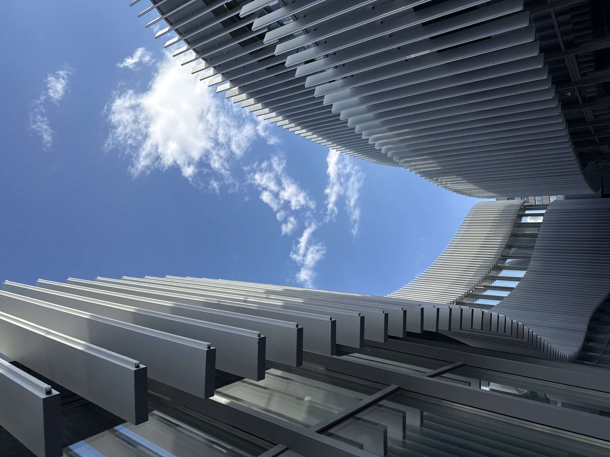 Low-angle shot of an architectural structure in Kazan, Russia. The view is upwards, capturing the sky and sections of two building facades. The facades are characterized by horizontal, gray, blade-like structures that create a layered effect. Between these structures, the blue sky with scattered white clouds is visible. Some of the building sections show windows. The architectural design has a modern and geometric aesthetic.