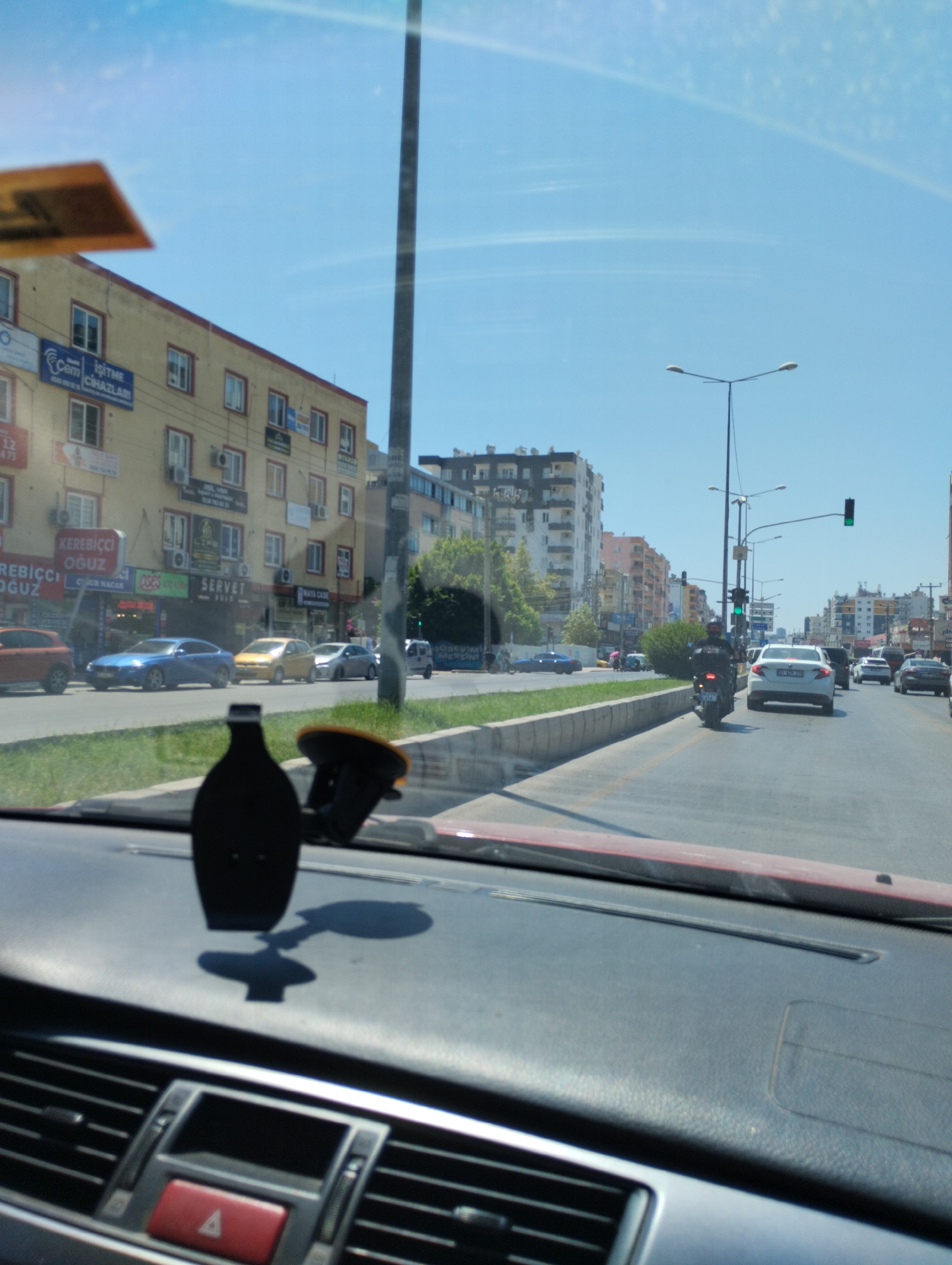 This is a photograph taken from inside a vehicle looking out at a city street in Mezitli, Turkey. The scene is viewed through the car's windshield, which shows some reflections. The street is lined with multi-story buildings, some with shops on the ground floor displaying signage. Cars and a motorcycle are visible on the road. The sky is clear and blue with some wispy clouds. The photograph captures a daytime urban environment with typical city traffic and architecture.