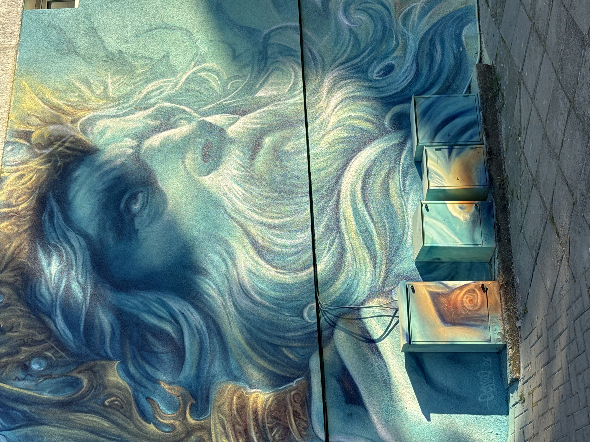 The image depicts a large mural on the exterior wall of a building in Burgas, Bulgaria. The mural features a detailed, stylized portrait of a bearded man with flowing hair, rendered in shades of blue, green, and gold. The man appears to be a mythical or divine figure, possibly Poseidon or a similar deity, with a crown or elaborate headdress. The mural covers a significant portion of the wall, extending from near ground level to the upper edge of the visible structure.
In the foreground, at the base of the wall, are three metal utility boxes. These boxes are also adorned with painted designs, complementing the style of the mural. The box on the left features a spiral design in orange and brown tones. The middle box displays a swirling pattern in blue and yellow, with what appears to be a stylized face or eye in the center. The rightmost box has blue and green swirling patterns.
The ground in front of the utility boxes and wall is paved with grey interlocking bricks, arranged in a basket-weave pattern. Sunlight casts distinct shadows from the utility boxes and a thin cable running horizontally across the lower part of the mural onto the brick pavement. The lighting suggests it is daytime, with the sun positioned to the left of the frame. The overall impression is one of urban art integrated with functional infrastructure.