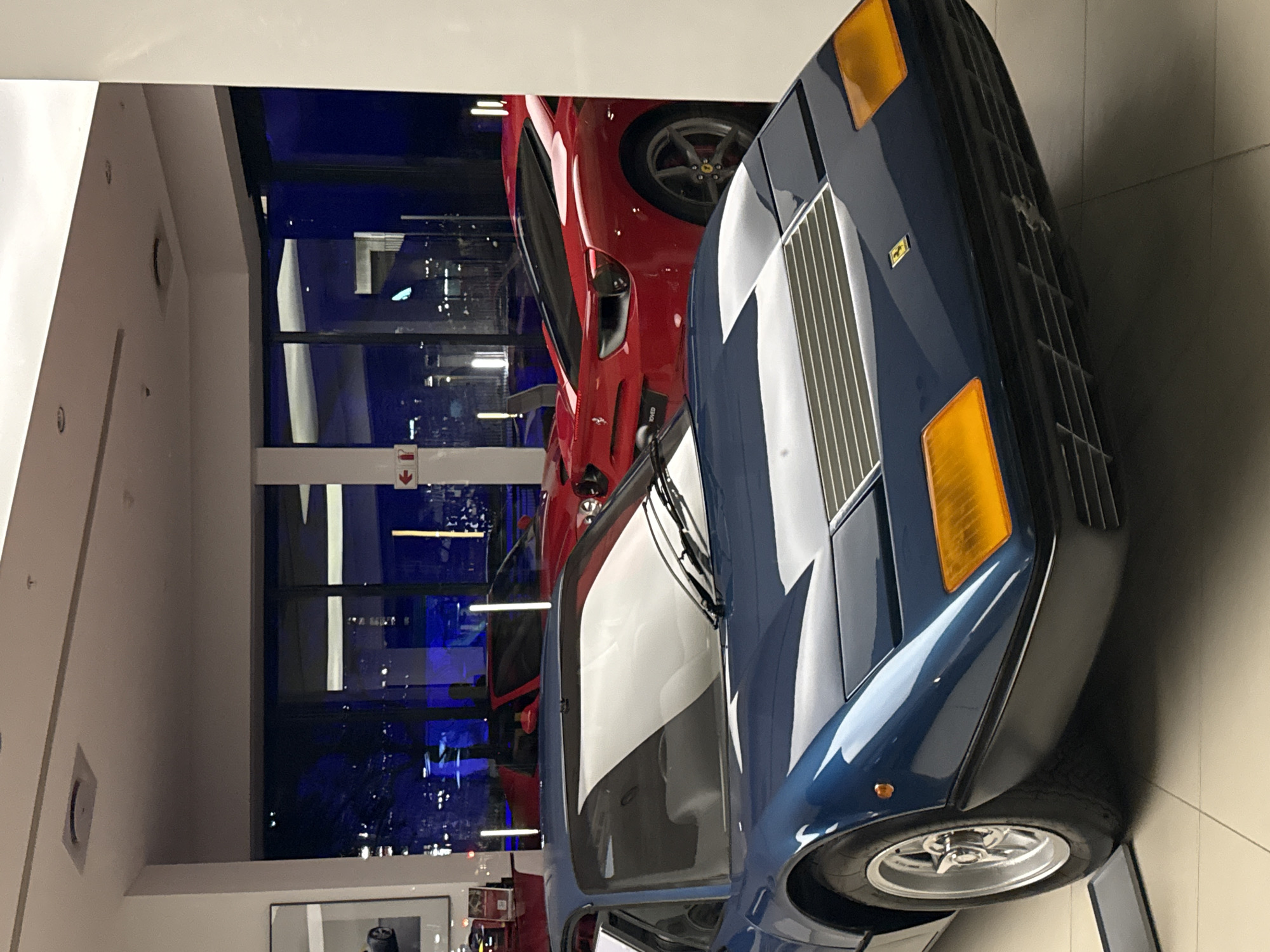 The image depicts the interior of a luxury automobile showroom located in the city of Cape Town, South Africa, at night. In the immediate foreground, a classic dark blue Ferrari Berlinetta Boxer (likely a 365 GT4 BB or 512 BB) is positioned, facing forward. Its pop-up headlights are retracted, revealing a horizontally slatted front grille and prominent amber rectangular indicator lights on the lower fascia. Silver multi-spoke wheels are visible on the front left. Behind this vehicle, through a large glass partition, a modern red Ferrari, identifiable as a 488 GTB or similar model, is displayed, oriented with its rear towards the viewer. An "Approved" label is partially visible near the rear of the red Ferrari. Another red vehicle is partially visible further back within the showroom. The interior environment features white walls, a light-colored tiled floor, and recessed overhead lighting. Through the large windows, the dark urban landscape of Cape Town is visible, illuminated by distant city lights and reflections of the showroom's interior. No individuals are present in the scene.