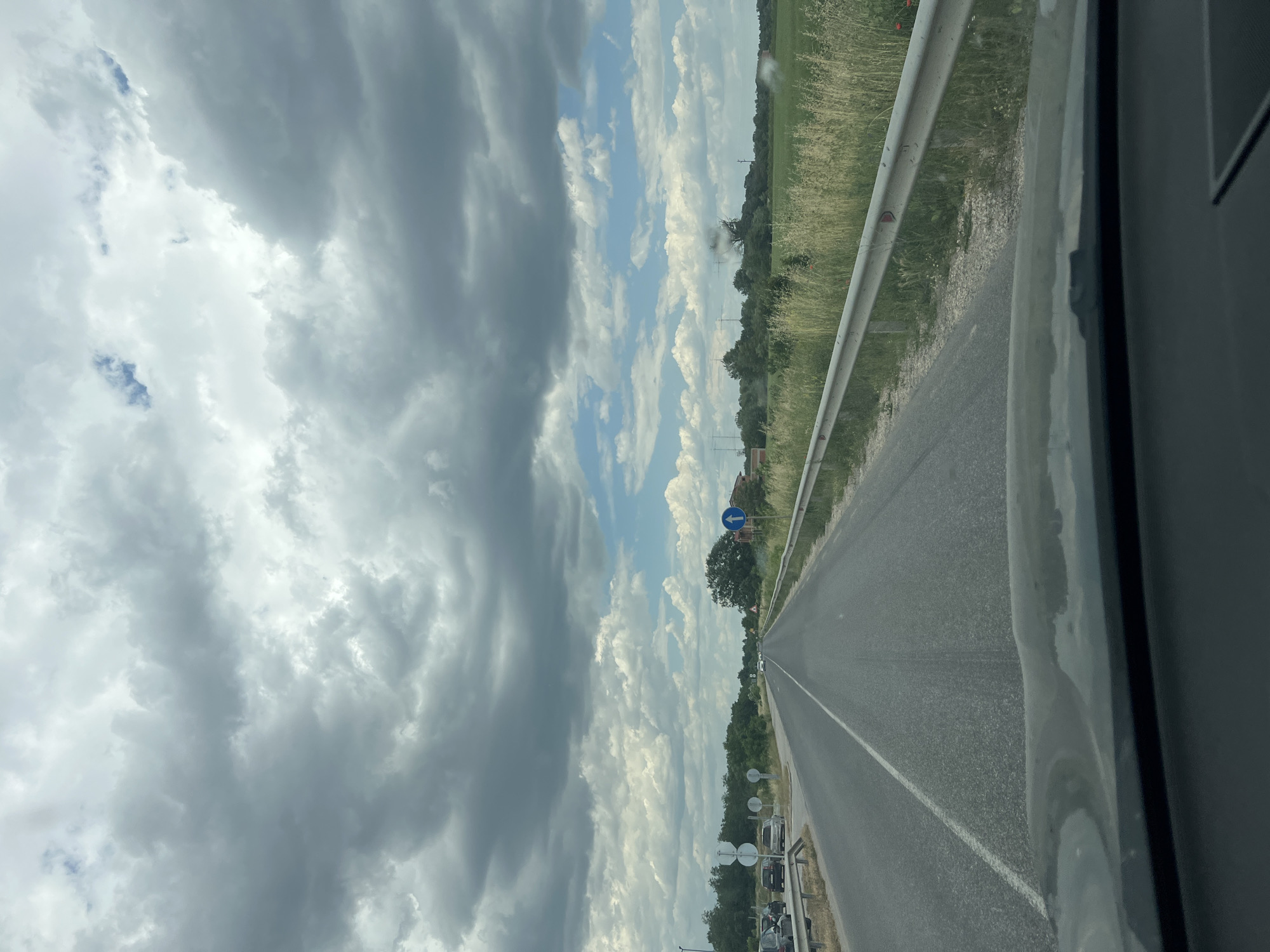 View from inside a vehicle traveling on a paved road in Kalekovets, Bulgaria. A metal guardrail borders the road on one side, with grassy fields and trees beyond. A blue and white road sign is visible. The sky is filled with clouds.