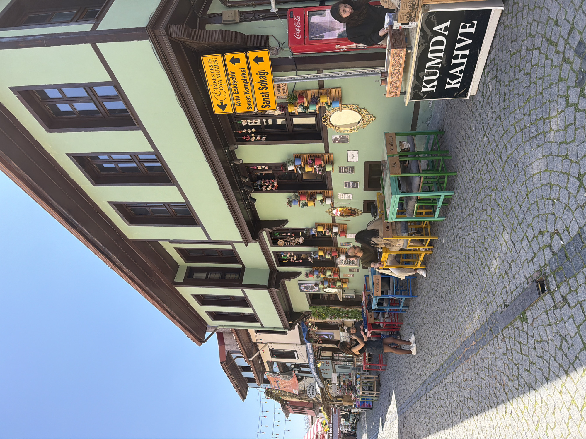 This photograph captures a vibrant street scene in Eskisehir, Turkey, under a clear blue sky. The main subject is a traditional building with light green walls and dark brown wooden beams, featuring numerous windows. Below this building, a lively outdoor cafe or market area unfolds on a cobblestone street.
Several people are present, including a woman standing behind a counter with a red Coca-Cola refrigerator, and others seated at colorful outdoor tables and chairs. One notable subject is a cat comfortably perched on a green chair. The street is paved with gray cobblestones, showing patterns of sunlight and shadow, indicative of a bright, sunny day.
The building walls are adorned with various decorative items, including small potted plants, ornamental mirrors, and framed pictures, creating an inviting and artistic atmosphere. Outdoor furniture, including tables and chairs in green, blue, yellow, and red, are arranged on the sidewalk.
Visible text includes a large black sign displaying "KUMDA KAHVE" (Sand Coffee). Yellow directional signs point to "IMREN ERSEN OYA MÜZESI" (İmren Erşen Embroidery Museum), "Avlu Eskişehir Sanat Kompleksi" (Courtyard Eskişehir Art Complex), and "Sanat Sokağı" (Art Street). A smaller sign indicates "ARİFBEY SOKAK". On the backs of the chairs, words such as "Sadakat" (Loyalty), "Aşk" (Love), "Mutluluk" (Happiness), and "Ölüm" (Death) are inscribed. In the background, signs for "SÜRMELİ KONAK" and "inci börek" are also visible. The overall setting suggests a bustling yet relaxed cultural and commercial street.