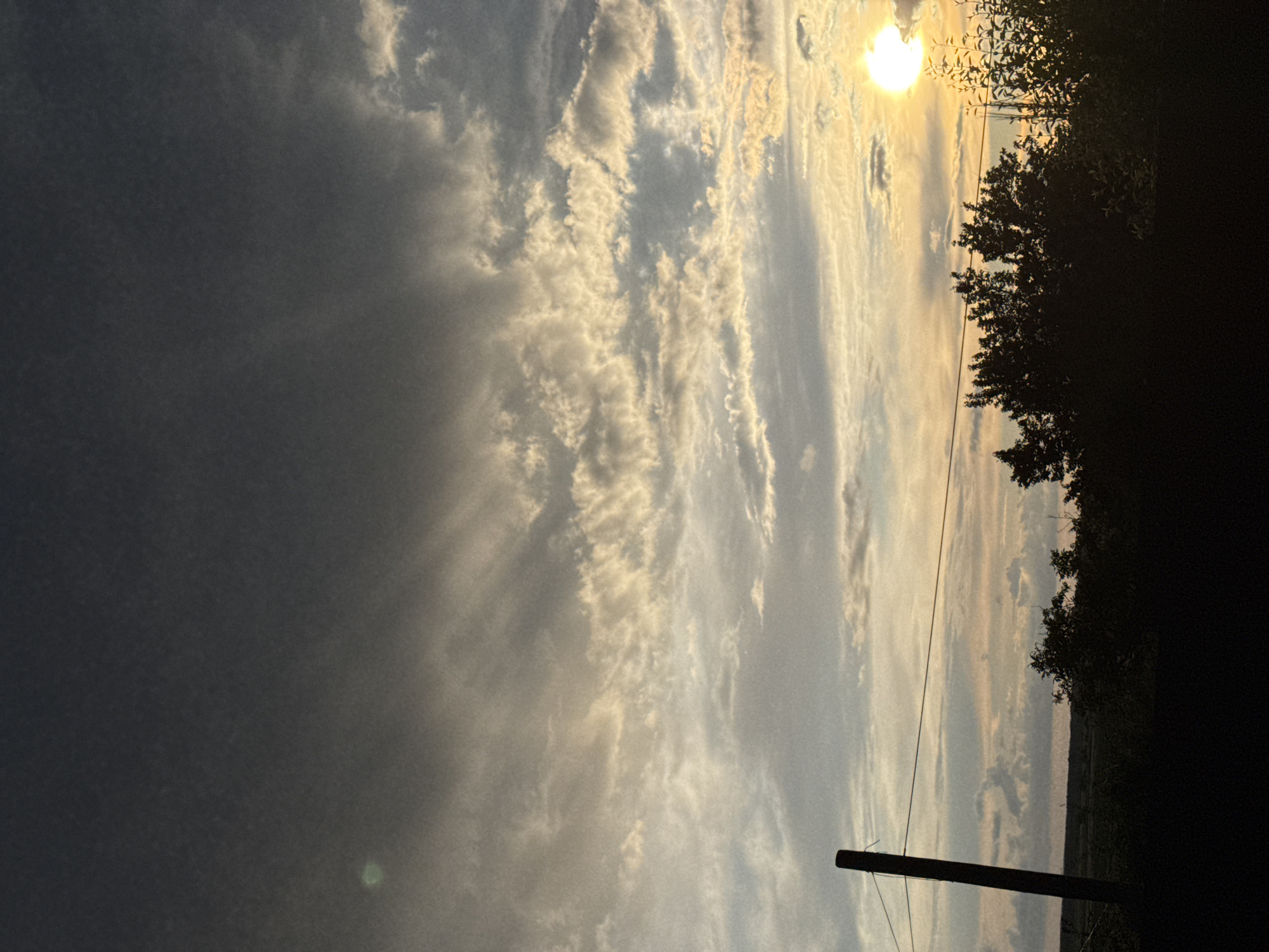 An outdoor, vertical, full-shot photograph captures a landscape scene of a cloudy sky at dusk. The sky is a mix of dark gray clouds and lighter, wispy clouds illuminated by the setting sun, which appears as a bright, round orb on the right side of the frame. In the foreground, a silhouetted line of trees and vegetation runs across the bottom of the image. A dark wooden post stands on the left side, with a wire strung horizontally across it. The location is described as Unknown, Unknown.