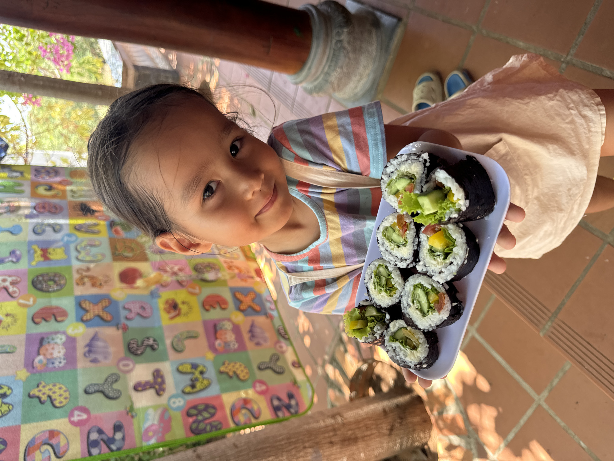 A young East Asian girl, appearing to be of pre-school or early elementary age, smiles brightly and looks directly at the camera. Her dark hair is pulled back, and she wears a multi-colored striped t-shirt in pastel shades (blue, yellow, pink, purple) and a light-colored, possibly beige or peach, skirt or dress. She holds a white rectangular plate with both hands, proudly presenting several pieces of vegetarian sushi rolls. The sushi, likely maki, features white rice, nori wraps, and visible fillings such as lettuce, cucumber, and yellow and red bell peppers or tomatoes.
The scene is an outdoor or semi-outdoor area, possibly a patio or veranda in Mũi Né, Vietnam, bathed in bright natural light, indicating it is daytime. The ground is covered with reddish-brown tiled flooring. A colorful children's playmat, adorned with various shapes, numbers (like "4" and "8"), and cartoon-like figures on different colored blocks, is spread on the floor to the girl's left. In the background, green foliage and pink flowering plants are visible, suggesting a garden setting, along with wooden structural elements and a decorative concrete base. A pair of light-colored shoes with blue accents can be seen on the tiled floor in the distance. The girl's cheerful expression conveys happiness and pride in presenting the sushi.