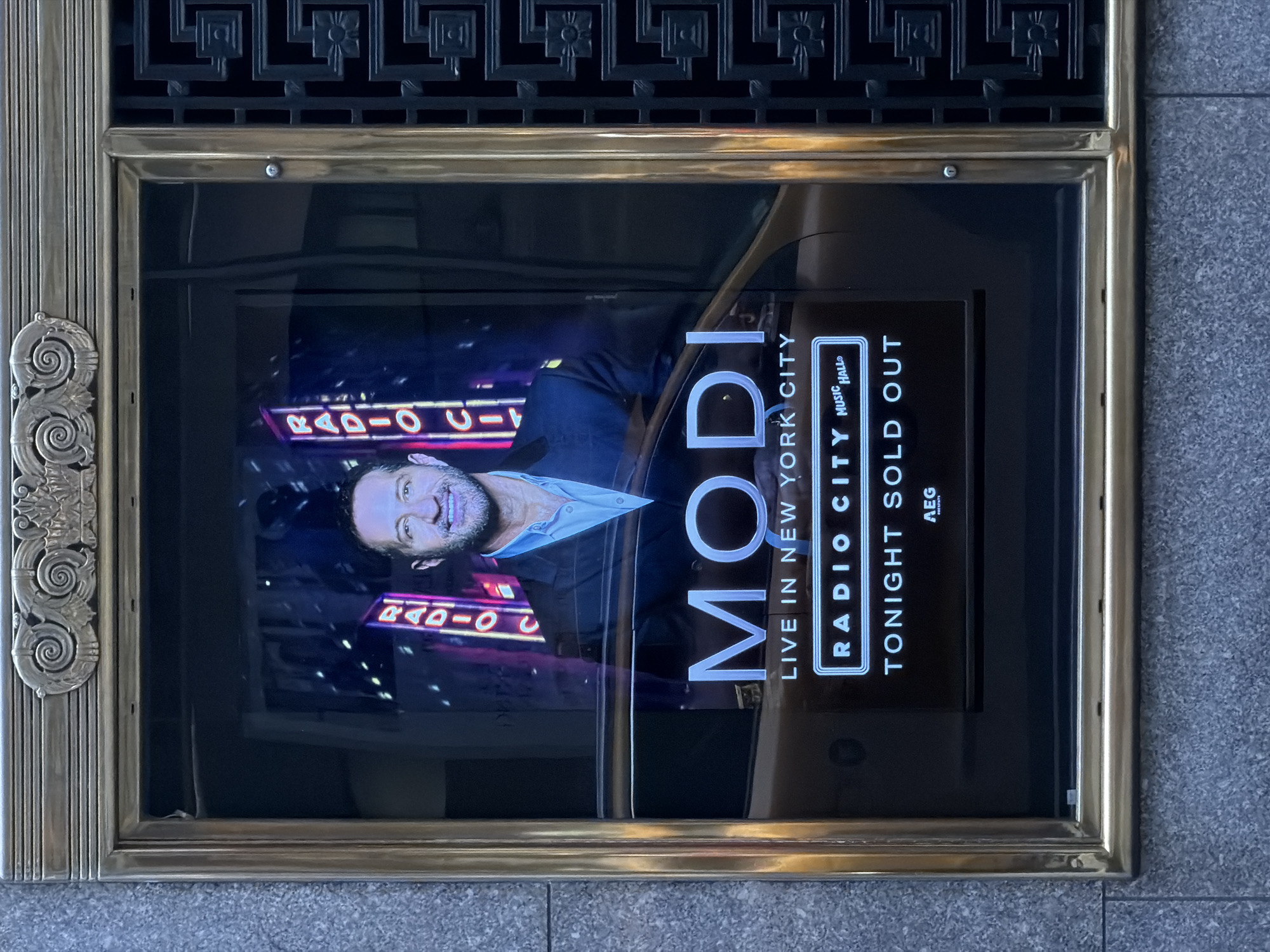 An upward-oriented photograph captures a display case set into a building exterior, featuring a digital advertisement for an event. The display case is framed by an ornate brass border, with decorative elements on the left side and a black decorative grille at the top. The surrounding building material appears to be light-colored stone or concrete paving.
The digital screen inside the case shows a man, presumably comedian Modi (Ahmed Al-Khabeeri), smiling broadly and looking towards the viewer. He has a beard and is wearing a light-colored collared shirt under a dark jacket. Behind him, neon signs partially visible spell out "RADIO CI" or "RADIO C!".
The primary text on the screen, oriented vertically, reads "MODI LIVE IN NEW YORK CITY". Below this, in a distinct rectangular box, is "RADIO CITY MUSIC HALL®". Beneath the venue name, the prominent message "TONIGHT SOLD OUT" is displayed, followed by the smaller text "AEG PRESENTS".
The overall impression is that of a successful event advertisement at a well-known venue in New York City. The time of day appears to be bright, based on the reflections and lighting on the brass frame and surrounding pavement, although the screen itself is brightly lit. No specific weather conditions are discernible.