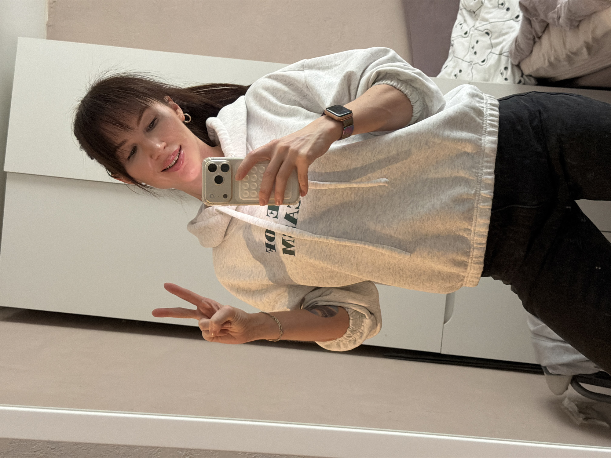 A woman takes a selfie in front of a mirror inside a residential space. She is wearing a light gray hooded sweatshirt, dark pants, and a wristwatch. She holds a smartphone in her right hand and makes a peace sign with her left. She has bangs and dark hair. The background includes a white cabinet and bedding. The location is in the city of Perm, Russia.