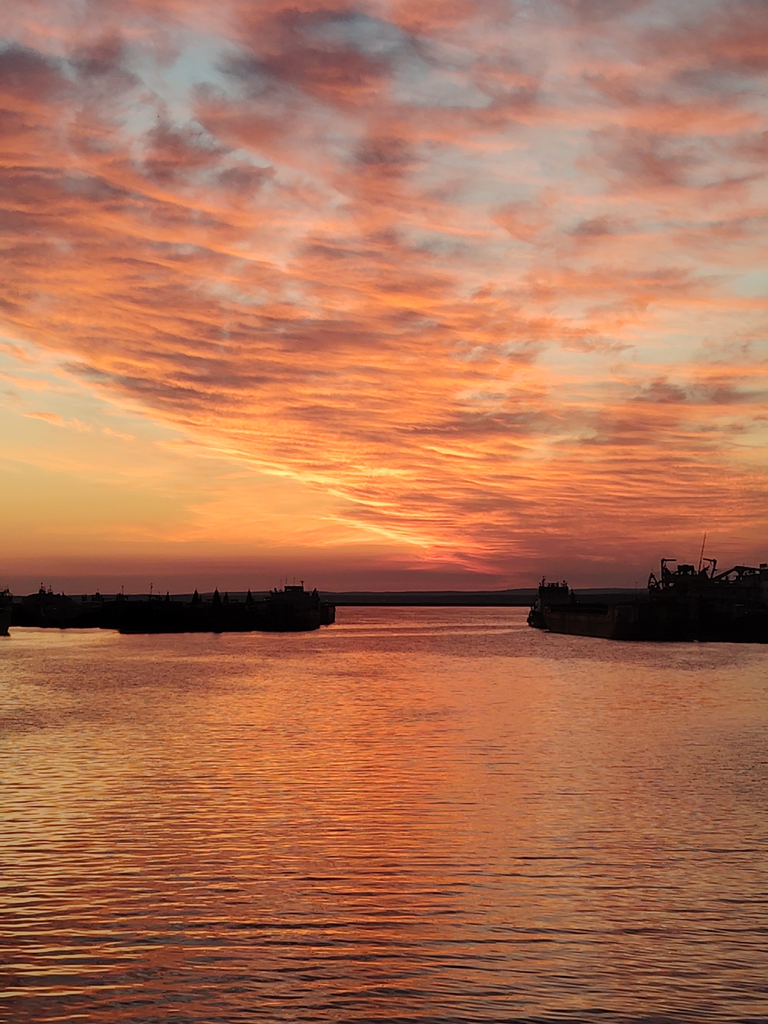 The image captures a vibrant sunset over a wide body of water, likely a river or reservoir, in Chaikovsky, Russia. The sky is dominated by dramatic, elongated clouds, painted in intense hues of orange, red, and pink, interspersed with patches of light blue-grey. The sun, though not directly visible, casts a bright, warm glow from just above the horizon, illuminating the clouds from below and creating a radiant band of yellow and orange light.
Below the colorful sky, the calm water reflects these fiery tones, appearing as a shimmering expanse of deep orange, reddish-brown, and golden light. The surface of the water is covered in numerous small, parallel ripples, creating a textured, almost striped pattern as it catches the low-angle light.
Along the distant shores, dark silhouettes of what appear to be several large industrial vessels or barges are visible. These ships are heavily shadowed against the bright backdrop, with their structures and superstructures outlined distinctly against the glowing sky and water. A faint line of distant land or shoreline separates the water from the lower part of the sky.
The scene conveys a sense of peaceful grandeur and the dramatic beauty of nature. There is no visible text in the image.