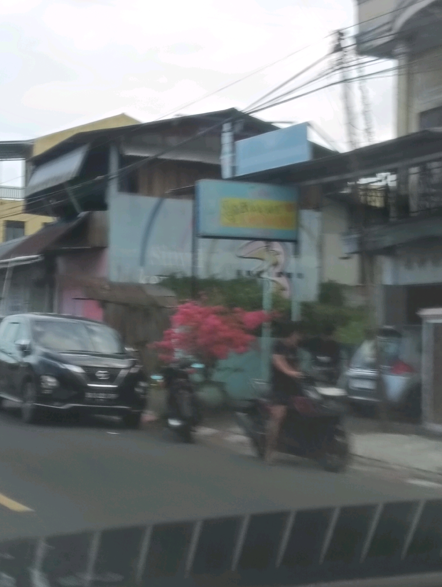 A blurred, street-level view captures a commercial area in Tomohon, Indonesia, under an overcast sky. In the foreground, a dark-colored SUV, possibly a Nissan, is parked on the asphalt road, its headlights illuminated. To the right of the SUV, a person is riding a dark-colored motorcycle, moving away from the camera. Behind the vehicles, a low-lying building with a yellow roof and dark eaves is visible. A pastel blue sign with illegible text is mounted above the building's facade, partially obscured by a darker sign. A cluster of bright pink flowers adds a splash of color to the scene, situated near the buildings. Electrical wires crisscross the sky, indicating an urban environment. The overall impression is one of a typical street scene in an Indonesian city, with commercial establishments and vehicular traffic.