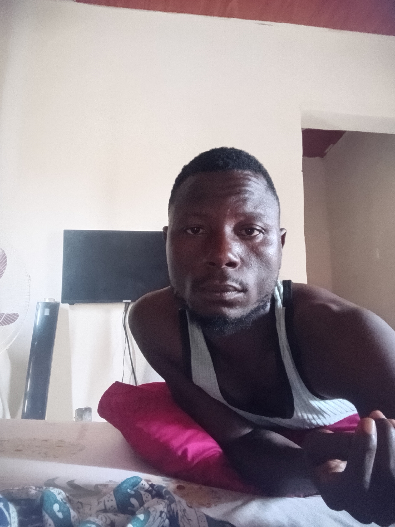 The image shows a close-up, front-facing selfie of a man with dark skin and short, dark hair. He is wearing a light-colored tank top and appears to be indoors, possibly in a bedroom. He is lying down, propped up on a bright pink pillow, with his left arm visible. In the background, a black flat-screen television is mounted on a white wall, and to the left, a white and silver fan is partially visible. The wall behind him is painted a light, neutral color. The overall lighting suggests an indoor setting with natural light possibly coming from the side. The scene is likely in Jalingo, Nigeria, given the context provided.