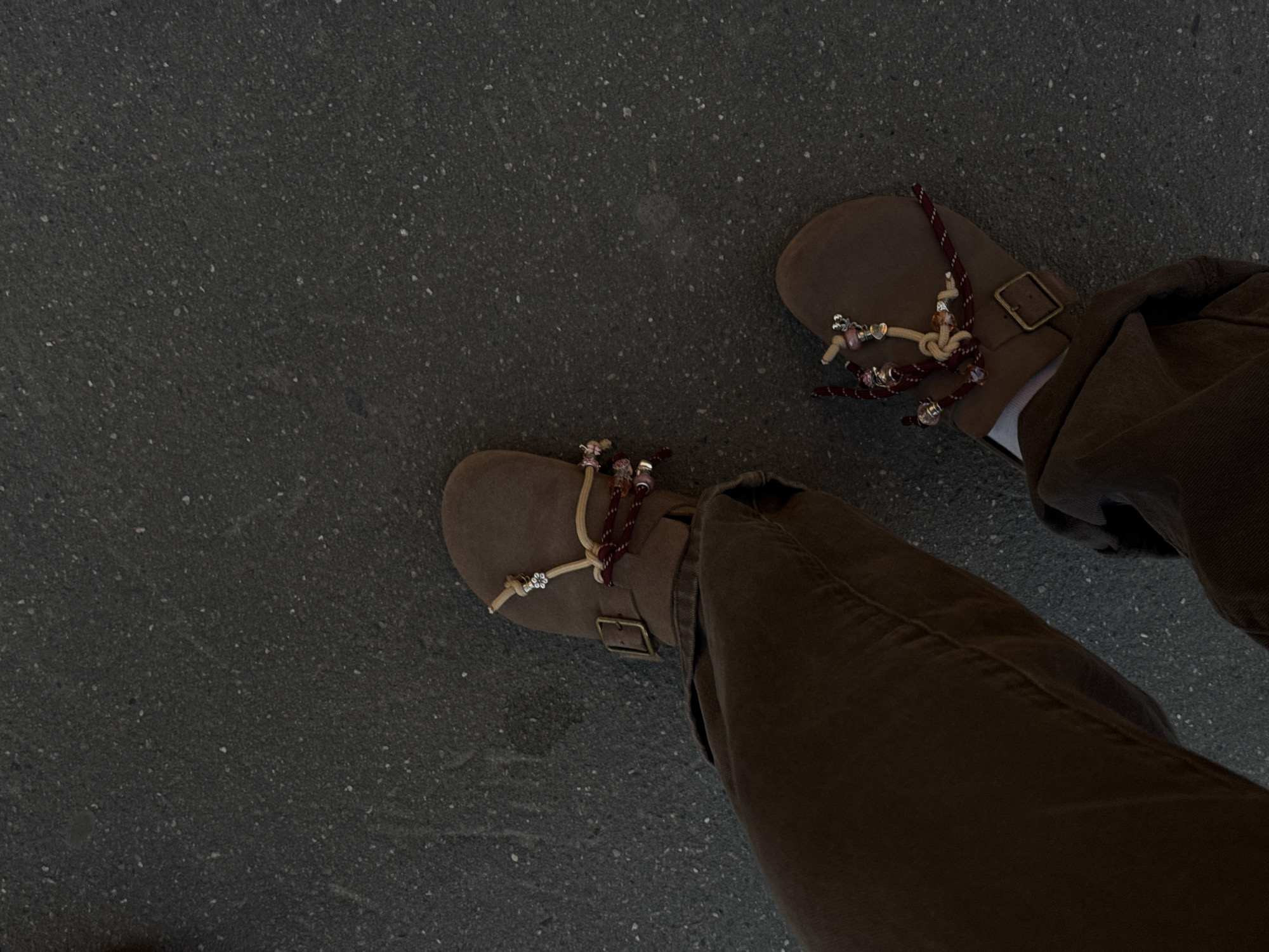 The image depicts a downward view of a person's feet and legs, clad in brown pants and brown, buckled sandals with decorative cords and beads, standing on a dark, gray concrete surface. The surface appears to be speckled with small, lighter-colored aggregates. The scene lacks any identifiable landmarks or indicators that would confirm a location in Moscow, Russia.
