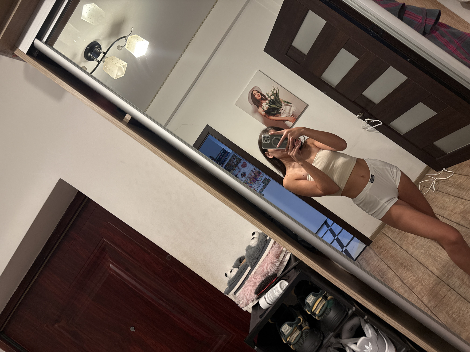 The image shows a young woman taking a mirror selfie in a room. She is wearing a white strapless top and white shorts. She is holding a smartphone in her right hand. The room appears to be a bedroom or dressing area, with a large mirror on a sliding door wardrobe, a dark wooden door with glass panels, a shoe rack with shoes and slippers, and a framed picture on the wall. The overall lighting suggests it is indoors during the day. The location of Chișinău, Moldova is not discernible from the image itself.