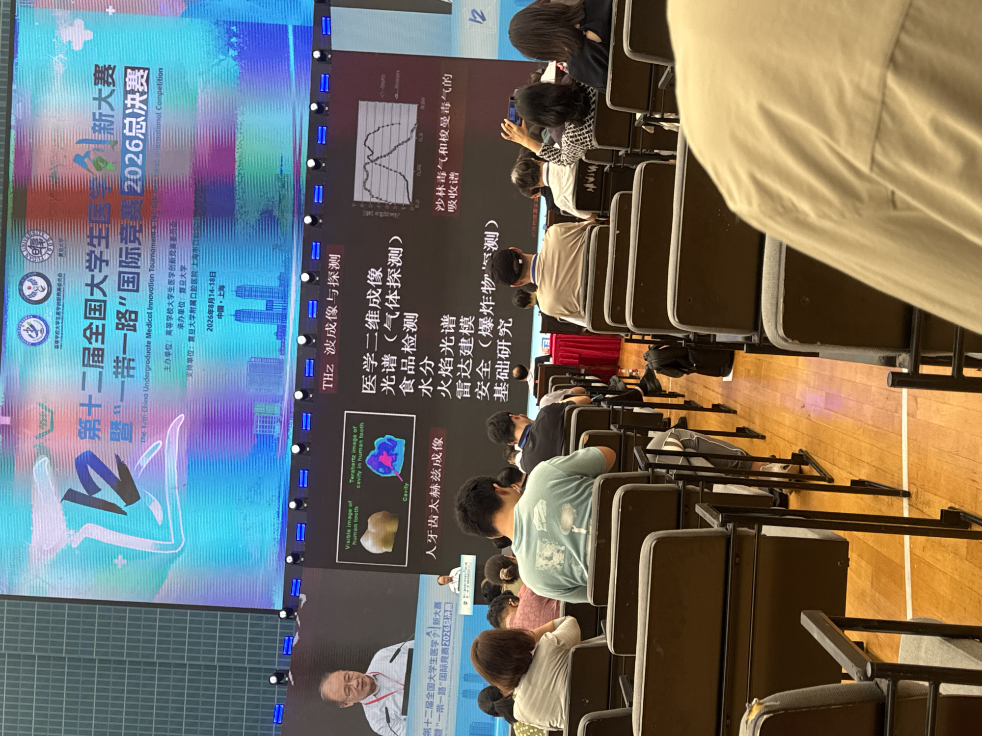 The image shows an indoor lecture hall in Yangpu District, China, likely during a conference or competition. A large screen displays information related to "The 12th China Undergraduate Medical Innovation Tournament - The Belt and Road International Competition 2026 Finals." The screen features text in both Chinese and English, alongside graphics of human teeth and spectral analysis data. A speaker is visible at a podium on the left side of the image, addressing an audience seated in rows of chairs. The audience members are diverse in age and gender, and some are engaged with their phones or listening attentively. The environment appears to be a modern educational or event space, with a wooden floor and overhead lighting.