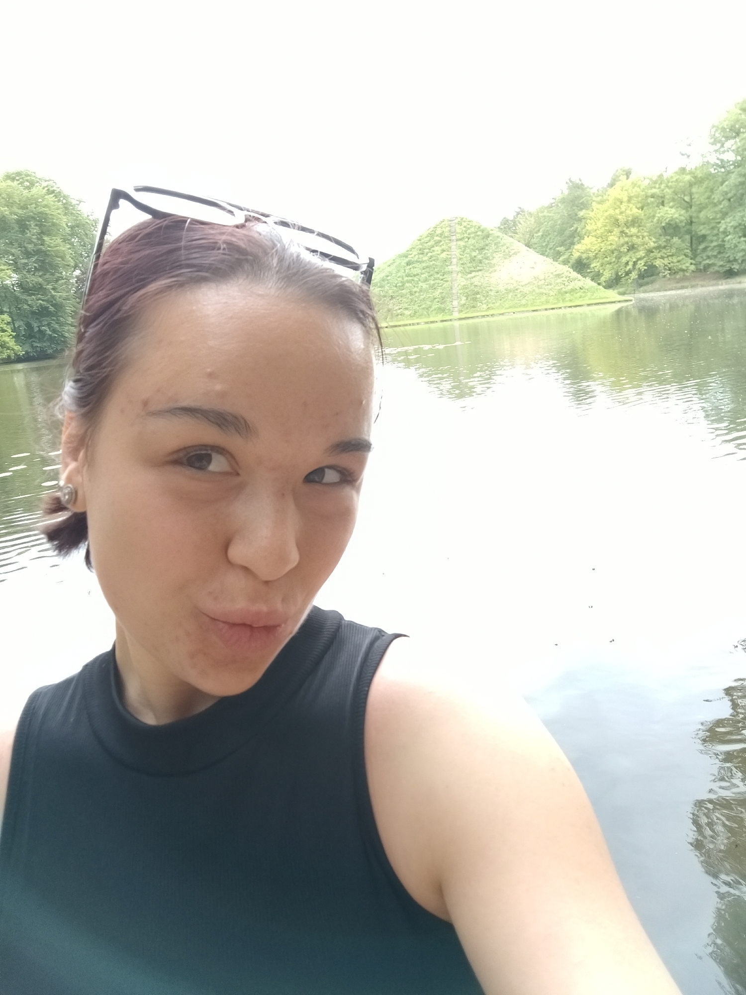 A young woman with dark hair and glasses on her head poses for a selfie in front of a body of water. Behind the water is a tiered, green pyramid. The woman is wearing a dark tank top. The photo was taken at the Seckenberg Pyramid in Bochum, Germany.