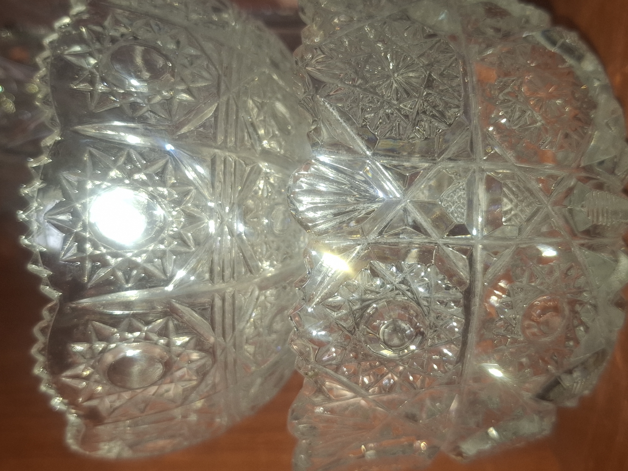 This image shows a close-up of two stacked crystal bowls with intricate geometric patterns and starburst designs. The lighting creates reflections and highlights on the faceted surfaces of the crystal. The background appears to be a warm, brown, possibly wooden surface. There are no people, animals, or any signs of human activity visible in the image. The image does not depict any specific location in Almaty, Kazakhstan, or any other identifiable geographical area.