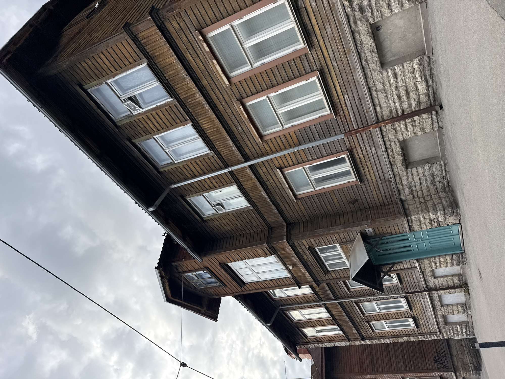 The image captures a tilted, low-angle view of a weathered wooden apartment building with a stone foundation in Tallinn, Estonia. The sky is overcast with visible clouds. The building features multiple windows with white frames and some are partially open. A prominent teal-colored door is set beneath a small, dark awning. A metal pipe runs vertically down the side of the building. The ground in front is paved asphalt. The architecture suggests an older residential district in Tallinn.