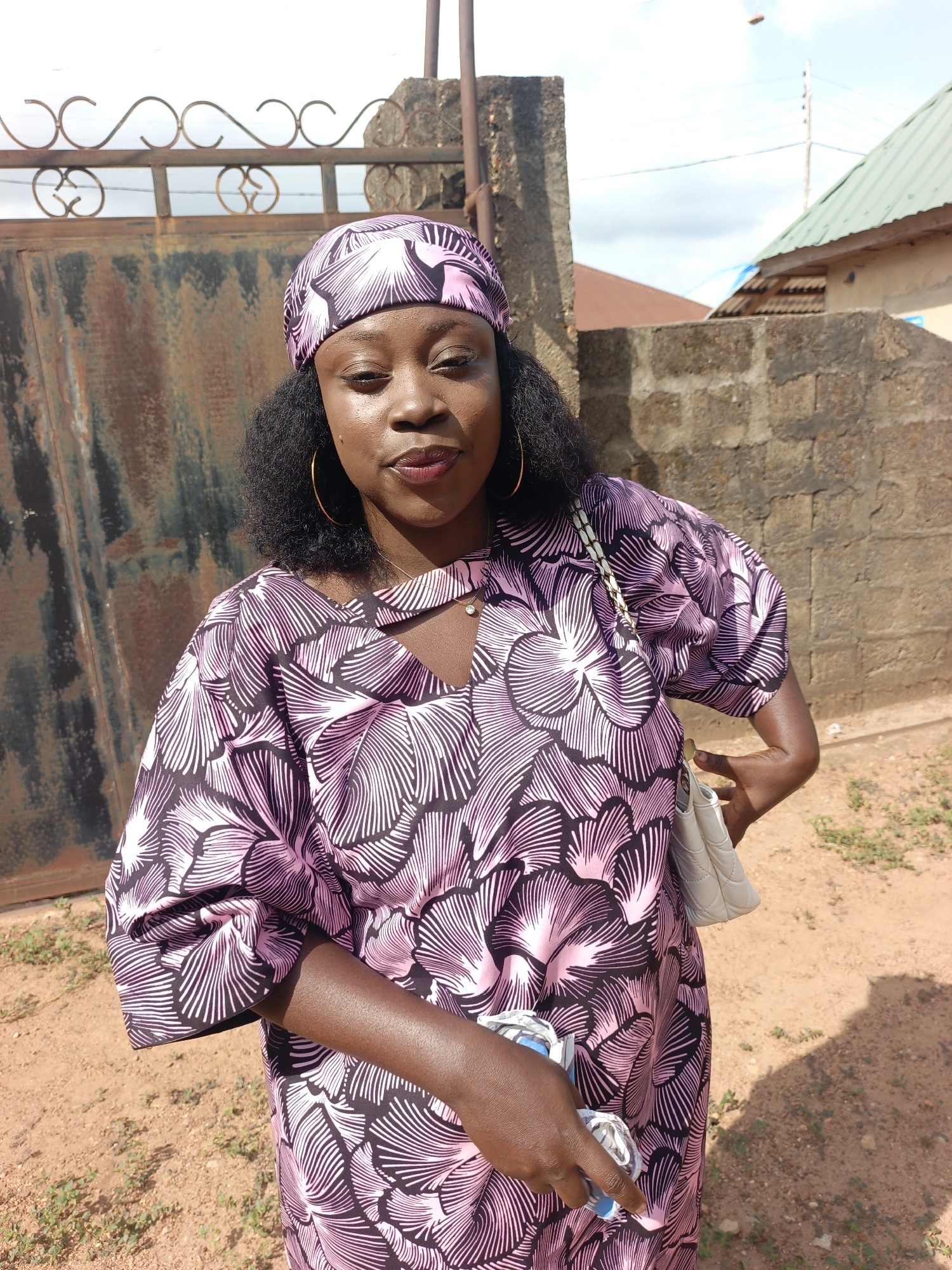 A young woman stands in front of a metal gate and a brick wall in what appears to be a residential area in Akwanga, Nigeria. She is wearing a pink and black patterned dress and a matching headwrap. She is also wearing gold hoop earrings and has a white purse slung over her shoulder. In her hand, she holds a small blue and white object. The environment is sunlit, with the ground covered in reddish soil and sparse vegetation. In the background, a building with a green roof is visible.