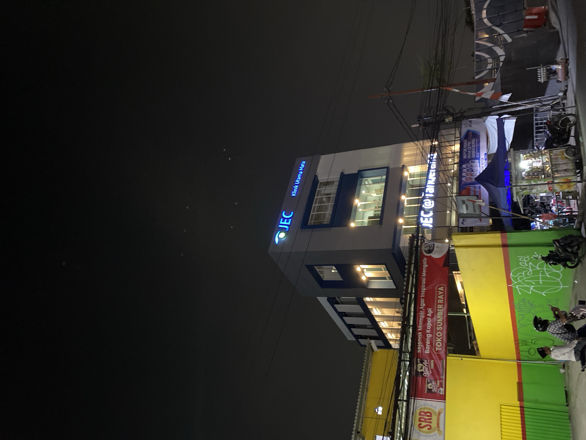 At night in Tangerang, Indonesia, a modern building with the illuminated sign "JEC Klinik Utama Mata" stands against a dark sky. The building features multiple stories with large windows, some of which are lit from within. To the left of the main building is a yellow structure with a red and white banner displaying "TOKO SUMBER RAYA" and other text. In front of the yellow structure, two young individuals, one wearing a white shirt and the other a checkered shirt with a helmet, are seated on a scooter. The foreground shows a street with various elements, including a food cart with a blue canopy, a motorcycle, electrical wires crisscrossing overhead, and some graffiti on a green and yellow wall. The scene captures a typical urban nighttime environment with commercial establishments and street life.