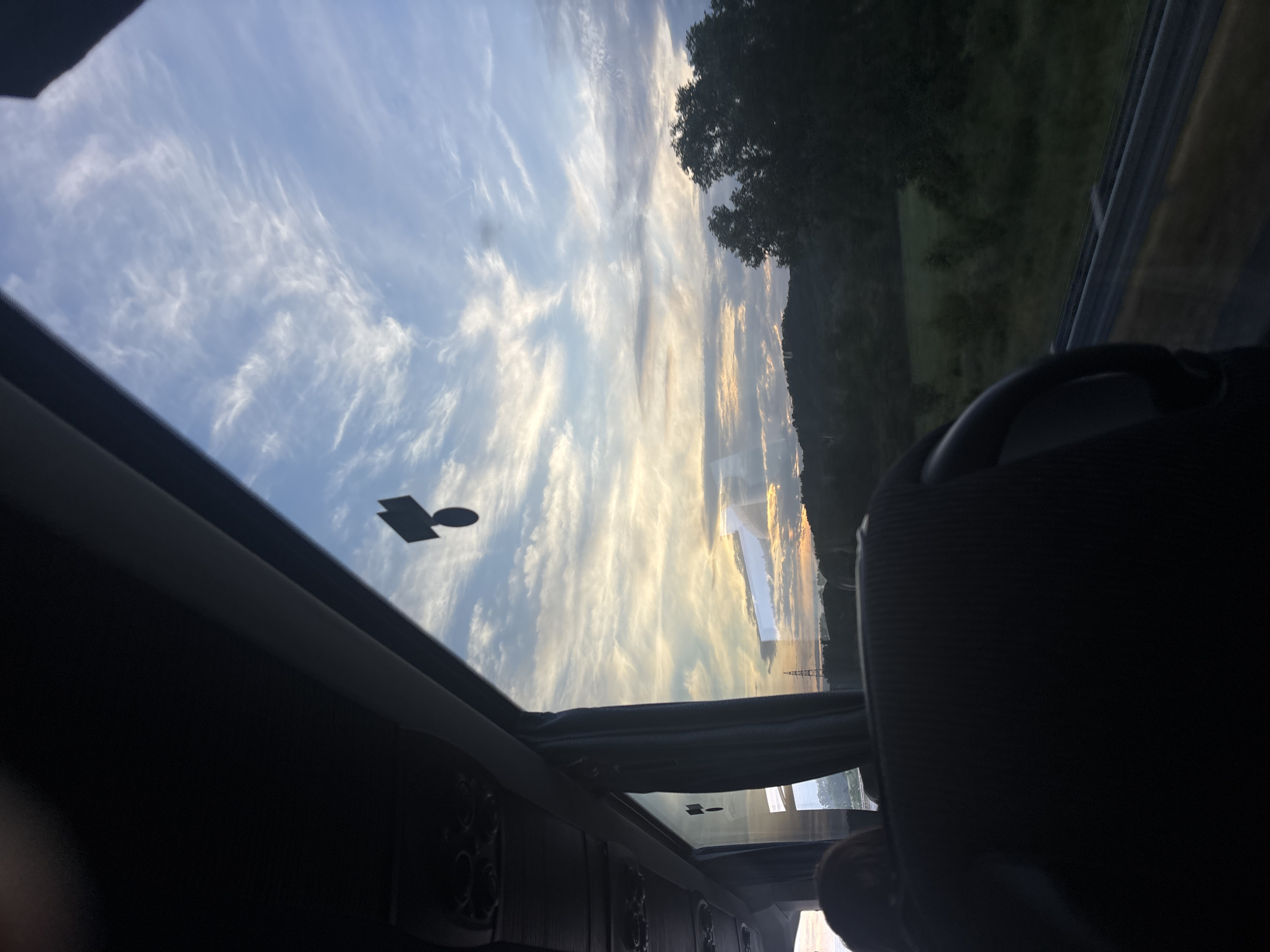 The image is an interior view of a vehicle, possibly a bus or van, looking through a sunroof. Above, the sky is visible, filled with wispy, cirrus-like clouds. Outside the vehicle, a landscape is partially visible, with greenery suggesting trees or hills. Power lines are seen through the sunroof. A small, round sticker with a rectangular addition is on the sunroof. Inside the vehicle, elements of the interior, such as seating and dashboard details, are seen. The image appears to be taken while the vehicle is in motion. The location is Obrenovac Urban Municipality, Serbia, deduced from the style of power lines and landscape.