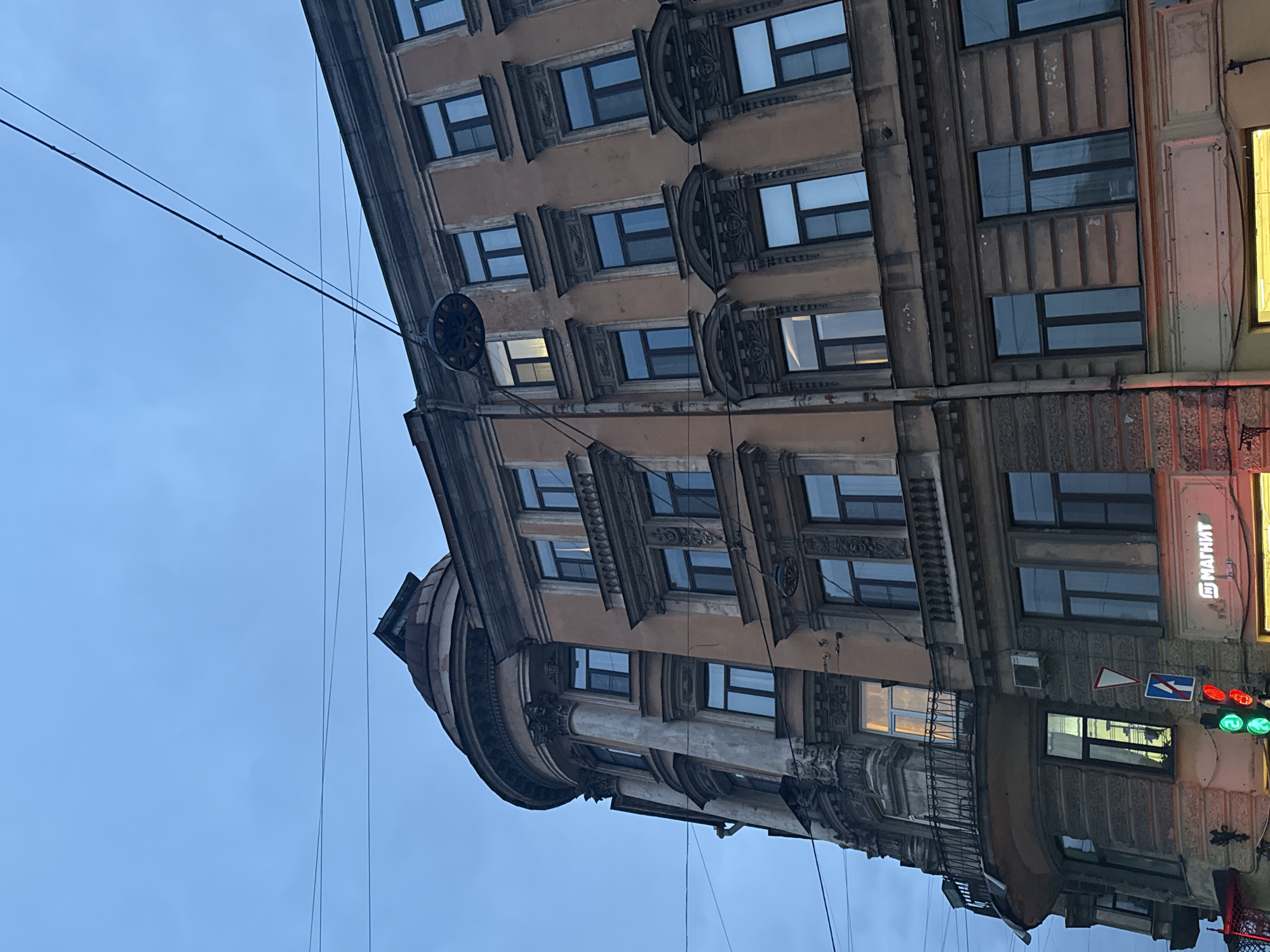 The image displays a multi-story building, characteristic of Saint Petersburg, Russia, under an overcast sky, likely at dusk or in the early evening. The building, constructed with a brownish-beige facade and dark, ornate architectural details, dominates the frame, viewed from a low angle. It features multiple rows of rectangular windows, many reflecting the dim sky or interior lights, with one window on the lower left showing a bright light from inside.
The architecture is classical, with elaborate decorative elements, including pilasters, cornices, and detailed window surrounds. The building has a distinctive rounded corner on the left side. Utility wires crisscross the blue-grey sky in front of the structure.
On the right side of the building, a brightly lit yellow sign is partially visible, suggesting a commercial establishment. Below it, a traffic light shows both red and green signals, and two street signs with white arrows on blue backgrounds are present. A prominent white sign with red Cyrillic text "МАГНИТ" (Magnit) is clearly visible, identifying a well-known Russian supermarket chain. A dark, circular, spoked object, possibly an old clock face or a decorative emblem, hangs from the building's facade near the top. The scene is an urban snapshot, focusing on the historical architecture and common city infrastructure.