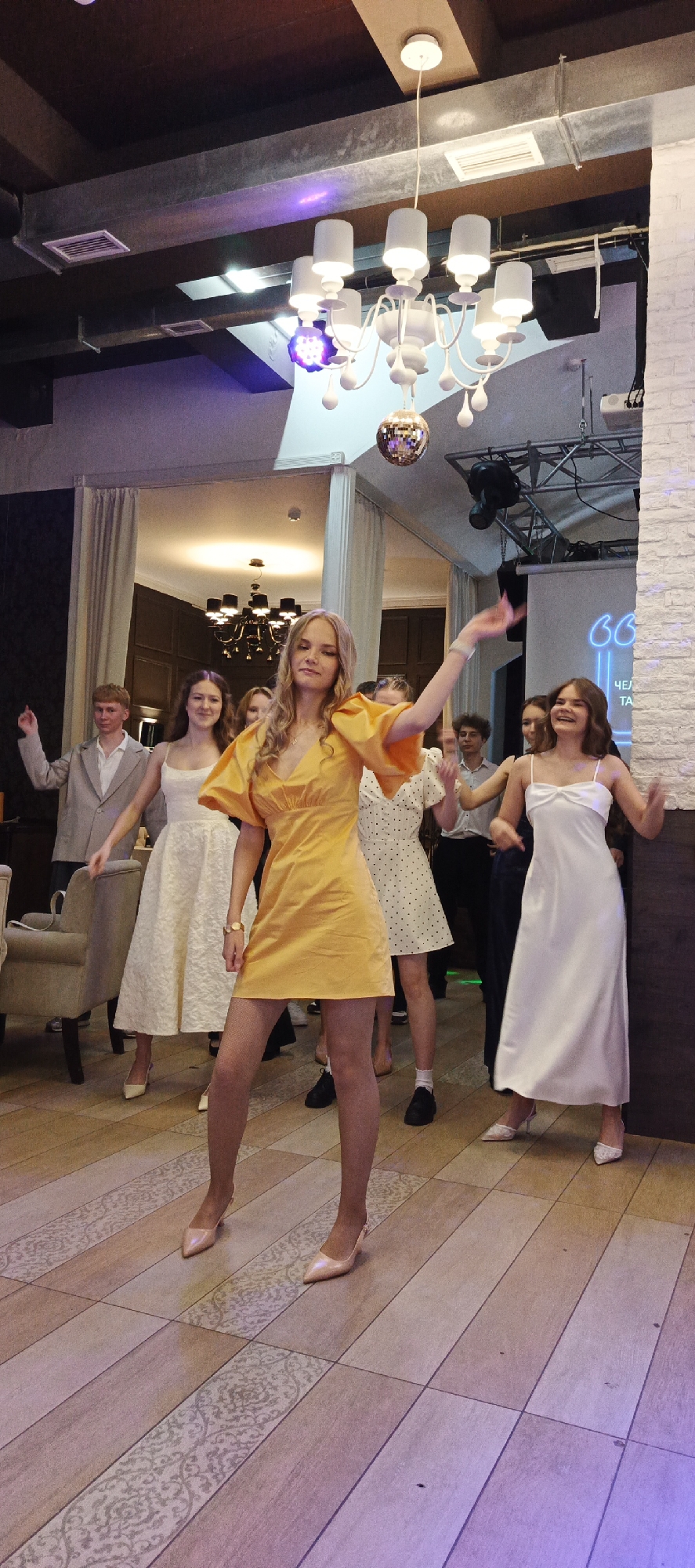 In an indoor venue, likely a banquet hall or restaurant in Chaikovsky, Russia, a group of young adults are seen dancing. The lighting is dim, with spotlights and a disco ball overhead creating a festive atmosphere. A woman in a bright yellow dress is the central figure, her arms outstretched as she dances. Several other individuals, some in formal wear like white dresses and suits, are also visible, either dancing or observing. The floor is made of polished wood tiles with decorative inlays. In the background, a projection screen displays text in Cyrillic, suggesting an event or celebration. The overall impression is one of a lively social gathering.