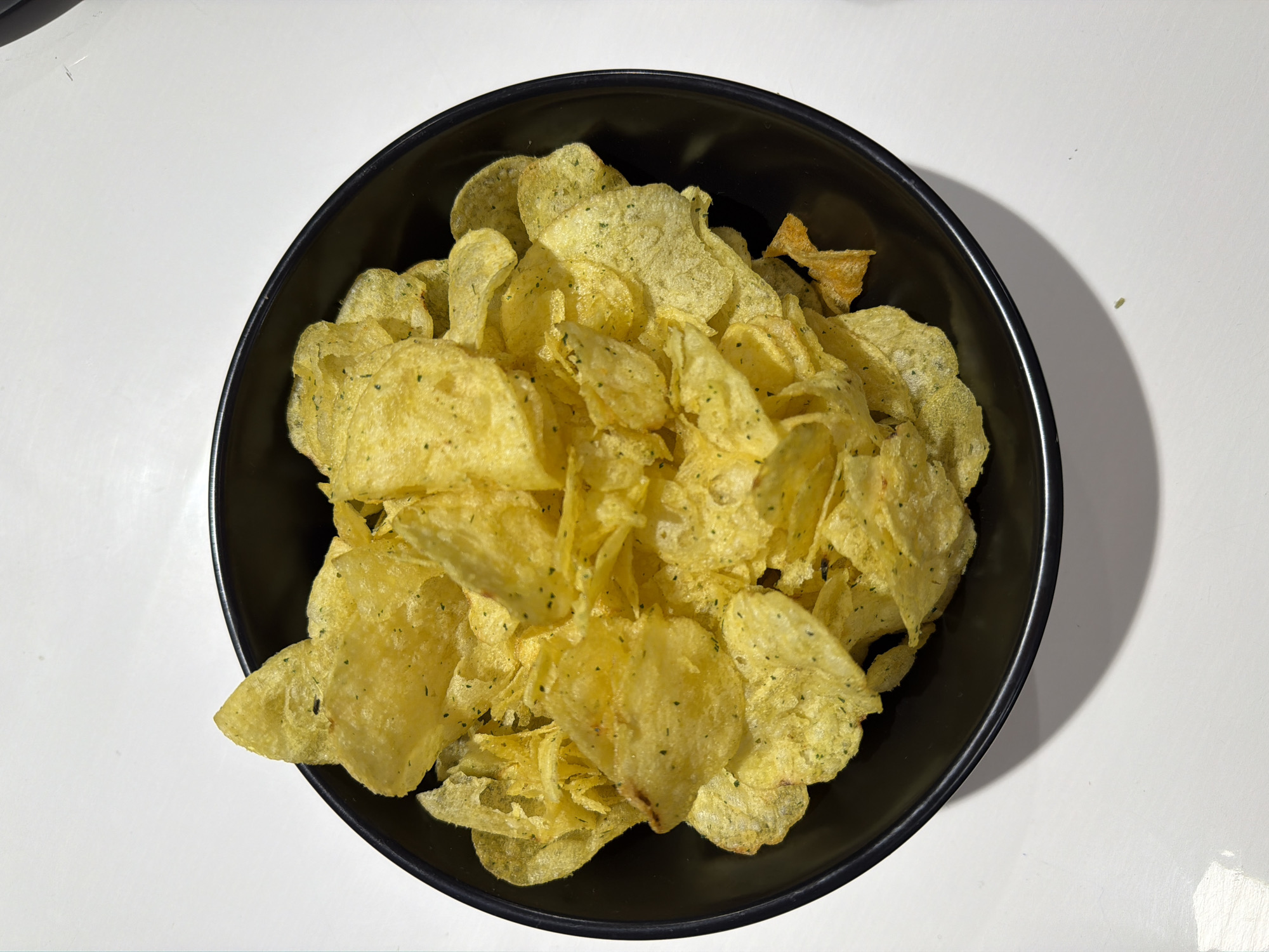 A black bowl filled with potato chips sits on a white surface. The chips are golden yellow and appear to be seasoned with herbs. The lighting casts a soft shadow to the left of the bowl. There are no people or any other objects visible in the image. The scene is indoors and appears to be a casual setting, possibly a kitchen or dining area. The image does not provide any information about the city of Buzuluk, Russia.