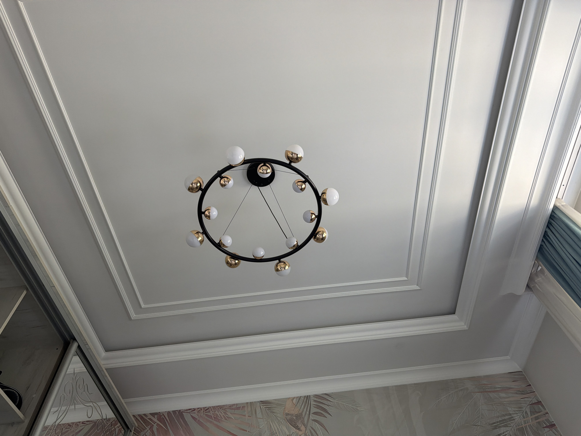 The image shows a circular chandelier with multiple white and gold globe lights hanging from a white, multi-tiered coffered ceiling. The chandelier is centrally located in the frame. The ceiling has ornate molding details. To the left, a portion of a wardrobe with a mirrored door is visible, reflecting a wall with tropical-themed wallpaper. To the right, light blue curtains are partially visible near a window. The scene appears to be an interior residential setting. There are no people or animals present in the image. The location is not specified as Джапалак, Kyrgyzstan.
