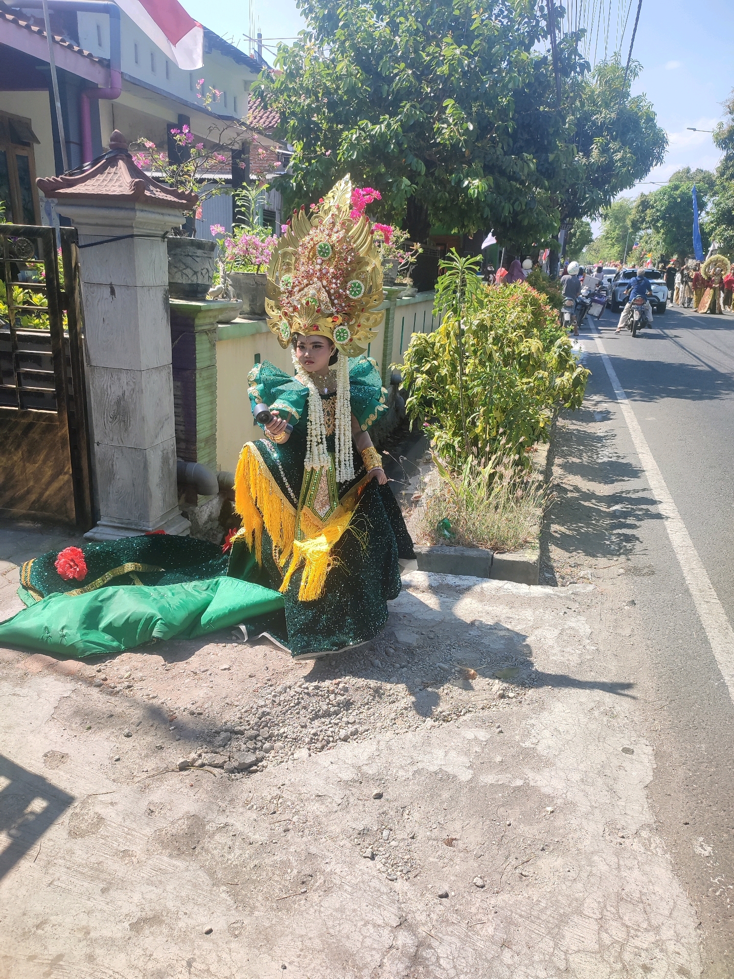 A young person dressed in an elaborate green and gold traditional costume, complete with a large, ornate headdress, is posed sitting on the ground next to a residential property's fence and gate. The costume features puffed sleeves, a beaded necklace, and a long, shimmering skirt adorned with yellow tassels. A large, draped green fabric with a red flower is spread out on the ground next to the person. Behind them, a street is visible with several motorcycles and people in traditional attire participating in what appears to be a parade or festival. The environment is sunny, with trees and bushes lining the street, and a building with a red and white flag is visible in the background. The scene is likely taking place in Nganjuk, Indonesia, given the cultural attire and the presence of a national flag.