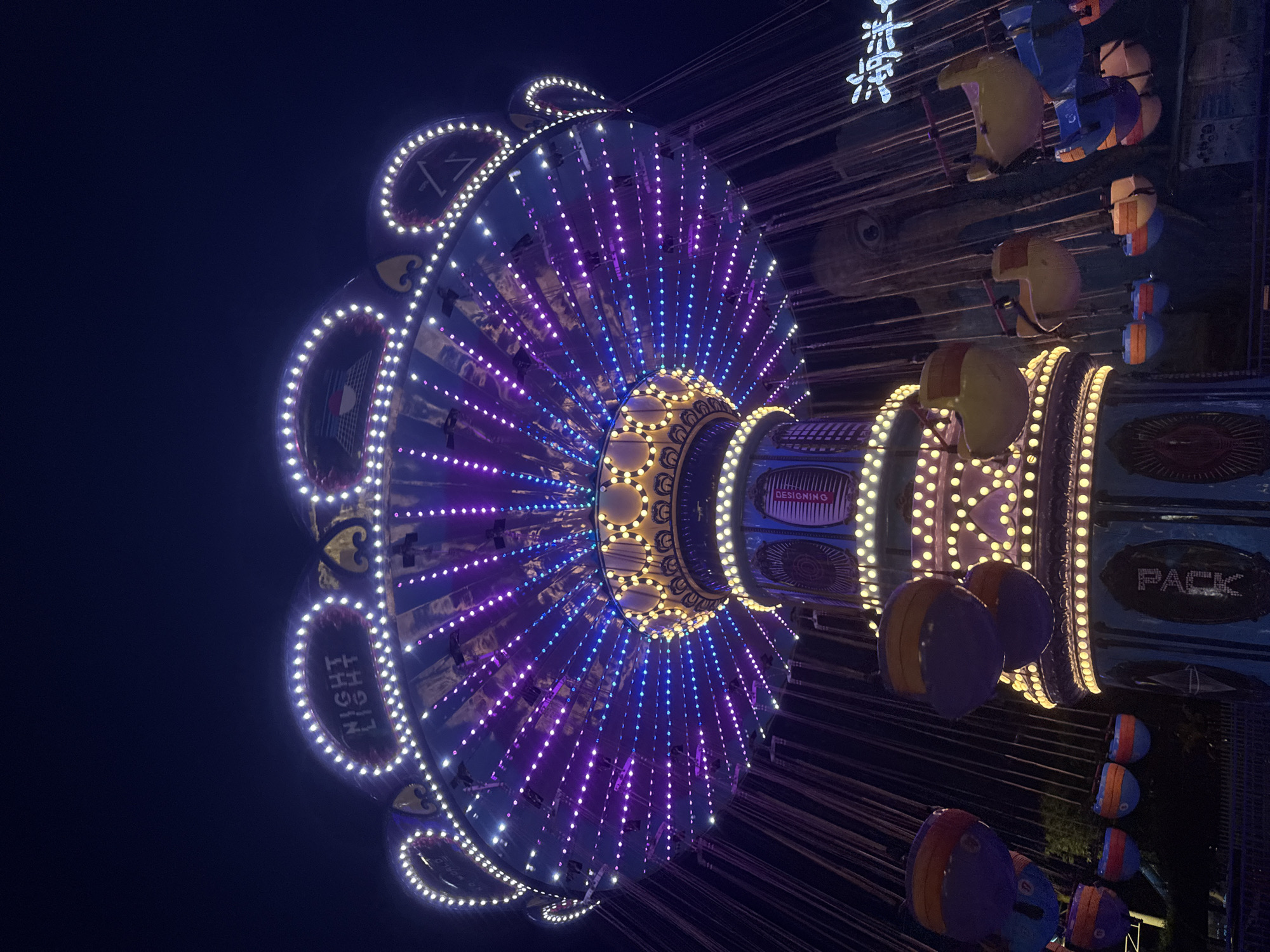 This is a low-angle, wide shot of a brightly lit amusement park ride at night. The ride, a classic swing carousel, is the central subject, illuminated by thousands of purple and white LED lights that form intricate patterns on its canopy and central column. The canopy flares out overhead, creating a circular, umbrella-like structure. Dangling from chains are colorful, bucket-style seats, some of which are visible in motion, suggesting the ride is operating. The background is dark, a deep blue-black night sky, with the silhouettes of trees and other park structures faintly visible. A neon sign with Chinese characters is visible on the right side of the image, indicating the location as Yiwu, China. The scene evokes a festive and exciting atmosphere, typical of an amusement park after dark.