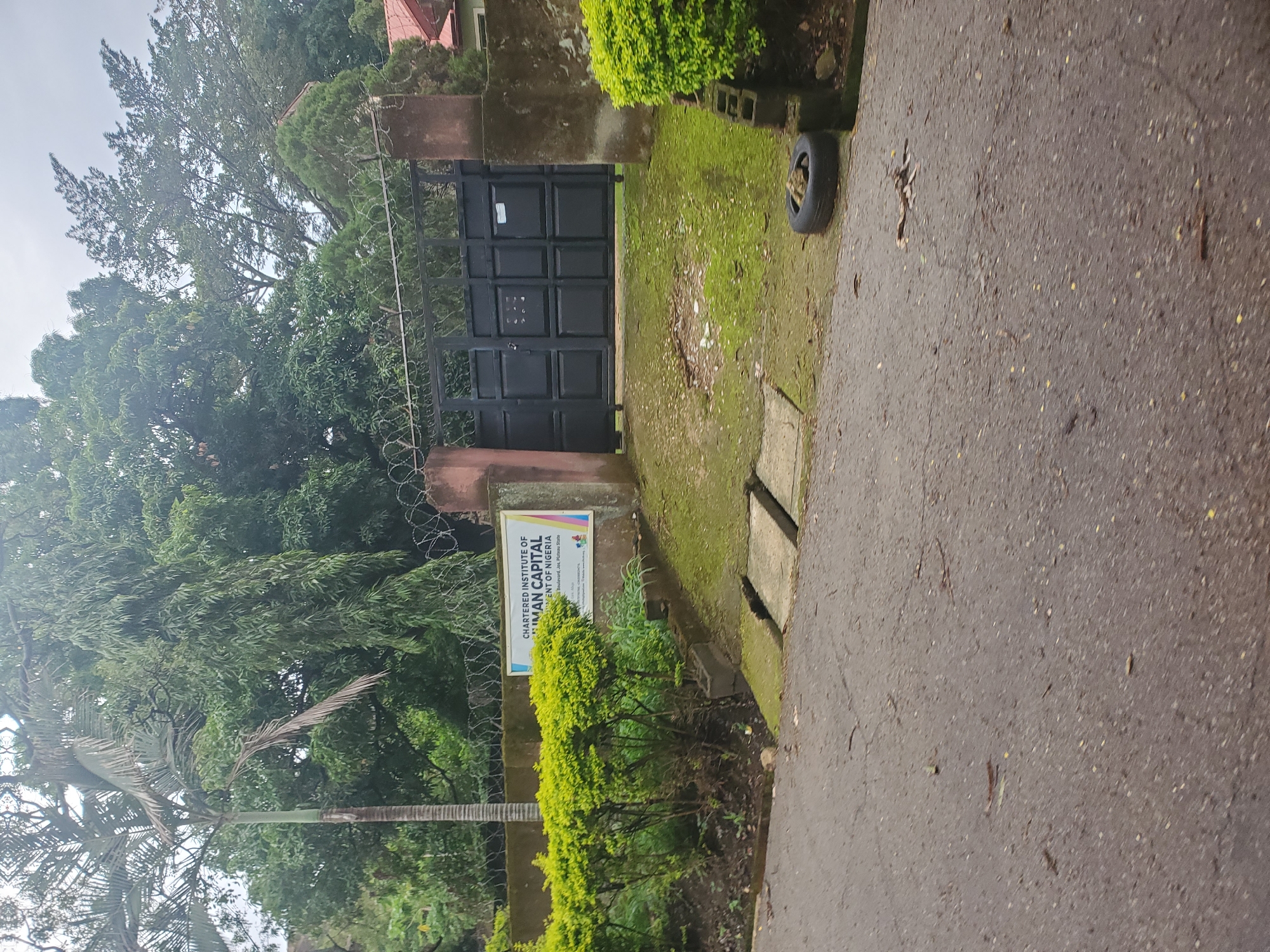The image shows the entrance to the Chartered Institute of Human Capital of Nigeria in Jos, Plateau State. A large, black metal gate dominates the center of the frame, flanked by two stone pillars. Above the gate, barbed wire is stretched across the top of a concrete wall, suggesting a security measure. To the left of the gate, a white sign with the institute's name and crest is mounted on the wall, surrounded by manicured green bushes. The ground in front of the gate is covered in grass and moss, with two concrete slabs forming a walkway. A discarded tire rests on the right side of the image, near a small pile of bricks. The overall scene depicts a well-maintained institutional entrance, hinting at a secure and organized environment.