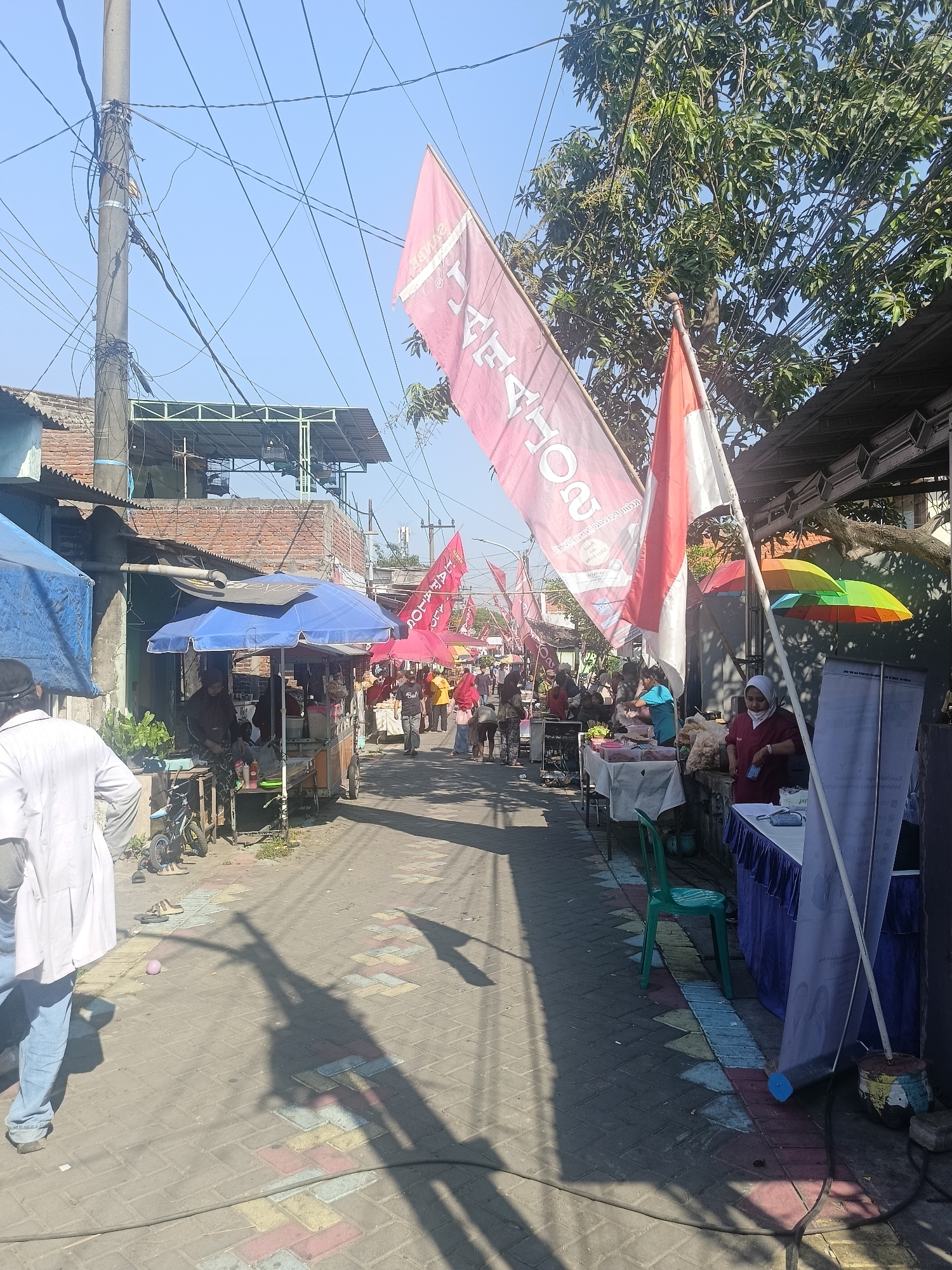 This image depicts an outdoor market scene in Gedangan, Indonesia, likely on a street or pathway. The environment is bright and sunny, with clear blue skies overhead. Power lines and a utility pole are prominent in the upper left, indicating an urban or semi-urban setting.
Several stalls or vendors are set up along the sides of the pathway, some with blue or red awnings. Various goods are displayed on tables. People are seen browsing, interacting with vendors, and walking through the market. One person in the foreground is wearing a white lab coat and blue jeans.
Decorative flags, including Indonesian national flags (red and white) and a large pink flag with text, are displayed, suggesting a festive or promotional event. Colorful umbrellas are also visible, one with red, yellow, and green sections. The pathway is paved with interlocking bricks in various colors. Shadows of people and overhead structures are cast on the ground. A few bicycles and small carts are present near the stalls. The overall impression is of a lively community gathering or a busy marketplace.