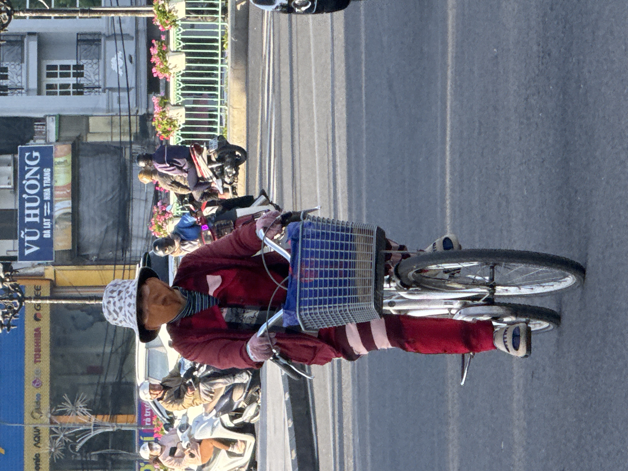 A person wearing a patterned bucket hat and a facial covering resembling a mask is riding a bicycle on a street in Da Lat, Vietnam. The person is dressed in a red jacket over a striped shirt and red pants with horizontal white stripes. They are wearing beige slip-on shoes with blue accents. The bicycle has a blue wire basket attached to the handlebars. In the background, several other people are riding motorcycles. One person in the foreground on a motorcycle wears a dark blue jacket and a helmet. Behind them, another person on a motorcycle wears a brown jacket and a white helmet. To the left, a woman wearing a pink jacket and a white helmet rides a white scooter. In the far background, buildings line the street, and a blue sign with white lettering reads "VŨ HƯƠNG DÀ LAT - NHA TRANG". Pink flowers in white planters are visible along a metal railing. The street is paved, and shadows are cast on the asphalt.