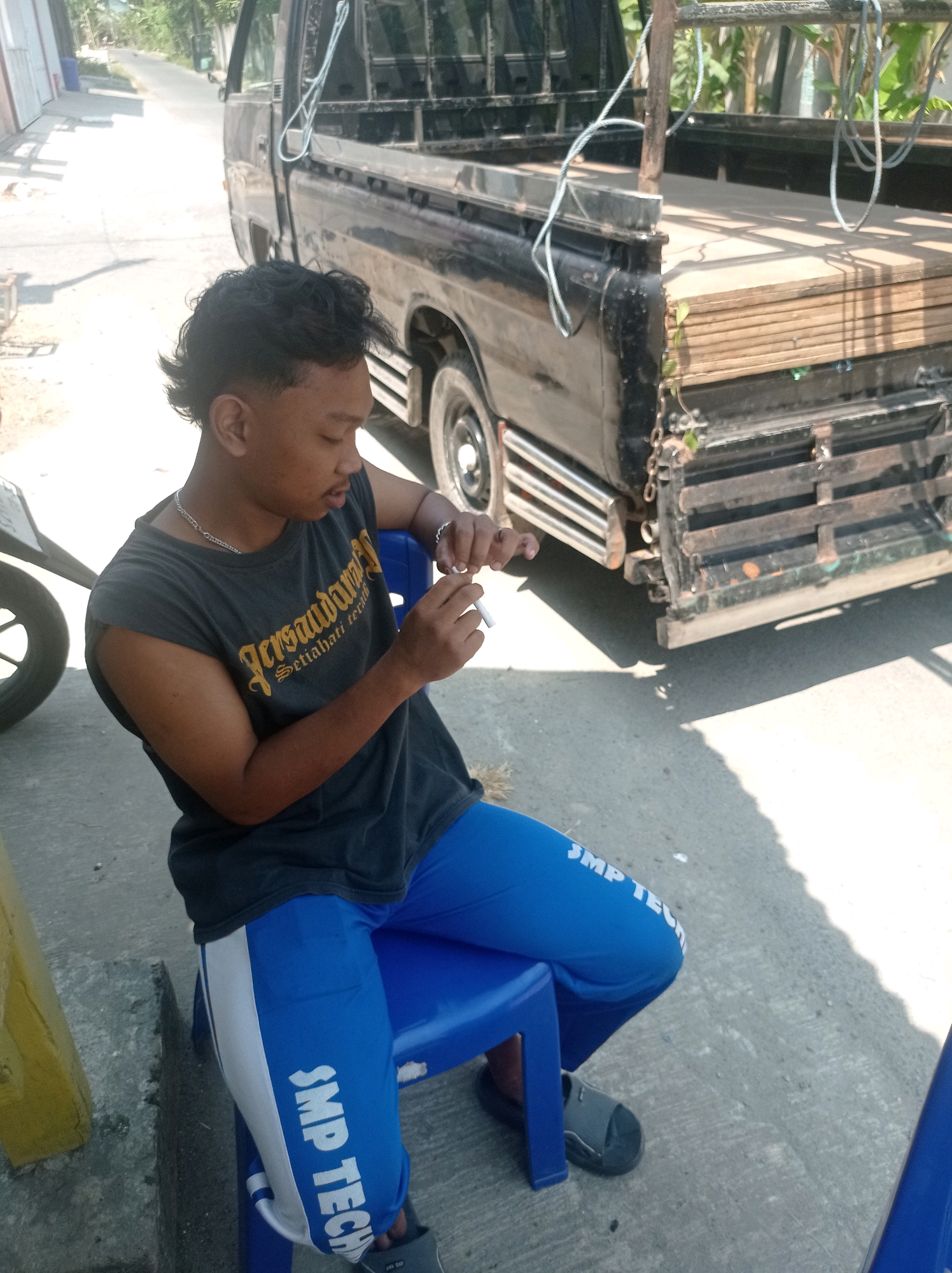 A young person with dark, styled hair, wearing a dark gray t-shirt and blue athletic pants, sits on a blue plastic chair in front of a black pickup truck. The t-shirt has yellow text that is partially visible. The pants have white stripes down the sides and white text that reads "SMP TECH". The person is holding a cigarette in their right hand and appears to be in the process of lighting or smoking it. The pickup truck is parked on an asphalt surface, likely a street or pathway in the city of Tuban, Indonesia, given the context. The truck's bed is loaded with stacks of wooden planks. Sunlight casts shadows on the ground. In the background, a street is visible, lined with trees and vegetation, and a building can be seen to the left. A portion of a motorcycle is visible on the far left of the frame.