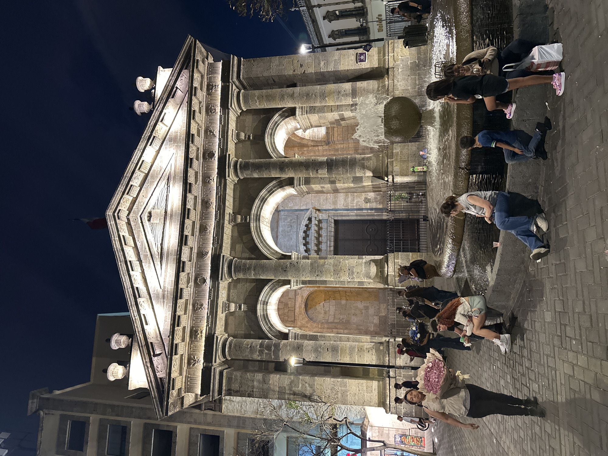 The image depicts the Guadalajara Cathedral in Guadalajara, Mexico, at night. The cathedral's facade is illuminated, showcasing its architectural details, including columns, arches, and decorative elements. A fountain is in front of the cathedral. Multiple people surround the fountain, interacting with it. One person is holding a bouquet of flowers. A street lamp provides additional illumination. The sky is dark, contrasting with the bright facade of the building. A building is visible in the background. The street is paved.