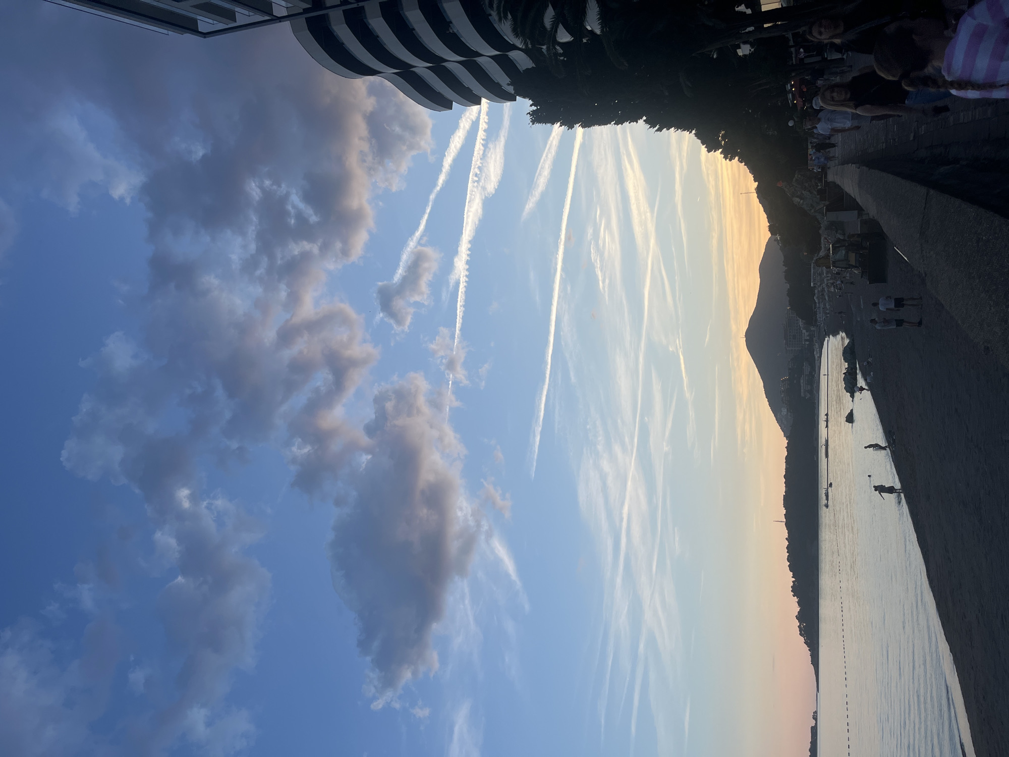 The image depicts a coastal scene in Bečići, Montenegro at dusk. The sky, a gradient of blue to orange near the horizon, is streaked with contrails and scattered clouds. A modern building with curved balconies is visible at the upper left, while the edge of a concrete promenade with people sitting and standing is present at the upper right. The sea occupies the lower portion of the image, reflecting the light. A dark, tree-covered mountain silhouette is visible in the background. People are seen on the beach and in the water.