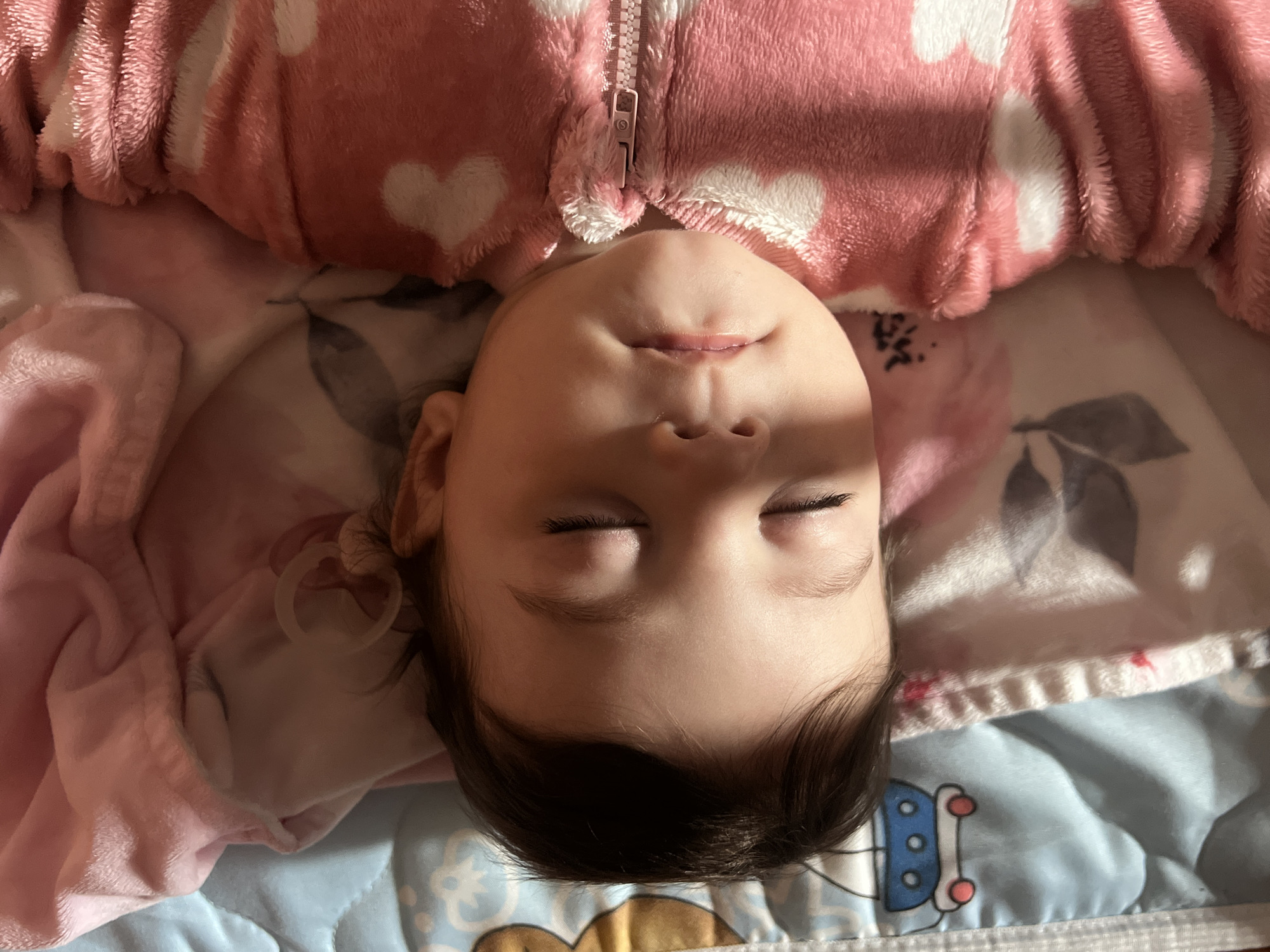 A baby lies on a blue and white patterned blanket and is wearing a pink and white heart-patterned fluffy jacket. The child's eyes are closed and they appear to be sleeping. A pacifier is near their ear. The photo is taken from an overhead angle. The location is not discernible from the image.