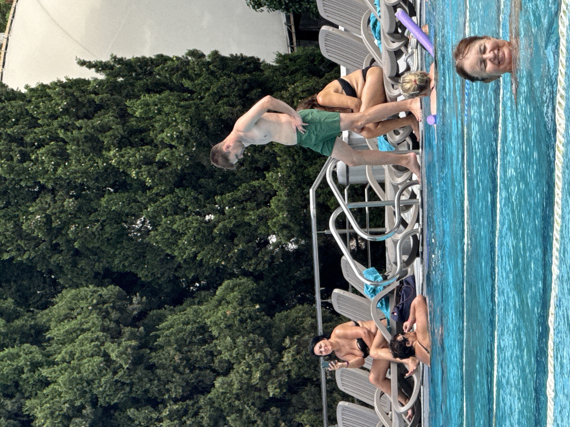 The image depicts a poolside scene in Yalta, Ukraine. In the foreground, a woman with brown hair is smiling in the water. In the middle ground, a man in green shorts stands, facing away from the camera, while a woman in a black bikini sits on a lounge chair. Another woman in a black bikini is also visible sitting on a lounge chair, holding a smartphone. Behind the lounge chairs, lush green trees line the background, with a white structure partially visible above the trees. The pool has lane markers and a purple pool noodle is present.