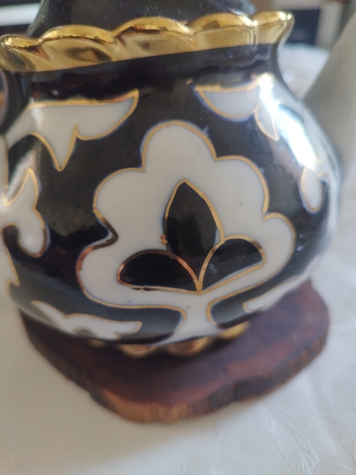 The image shows a close-up of a decorative ceramic teapot with a black and white floral pattern outlined in gold. The teapot has a fluted gold rim and rests on a dark wooden base. The background is a white, textured surface, likely a tablecloth. The scene appears to be indoors, with soft lighting. There are no people or animals visible in the image. The location provided, Чистенское сельское поселение, Ukraine, is noted as a rural settlement.