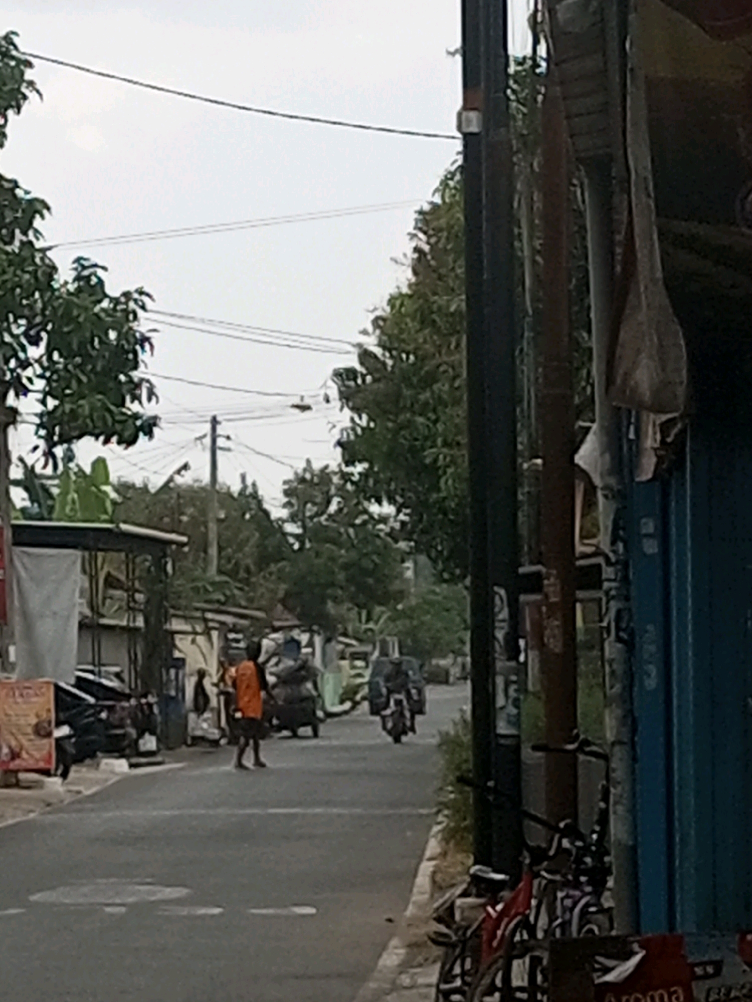 The image depicts a street scene in Kalikidang, Indonesia. A man wearing an orange shirt and dark shorts walks across the road. Several motorcycles are parked on the left side of the street, and a motorcycle is visible in motion in the background. A three-wheeled vehicle, possibly a rickshaw, is also present. The street is paved with white dashed lines marking lanes. Trees line the sides of the street, and utility poles with numerous wires are visible. Buildings and structures, some with open fronts, are situated along the road. The sky is overcast. In the foreground, several bicycles are parked near a building with a blue facade.