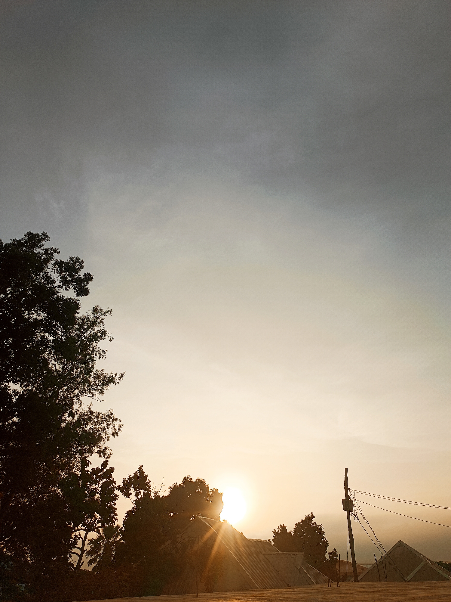 The photograph captures a scene at sunset in Bukuru, Nigeria. The top portion of the image shows a sky with gradient from grey to yellow due to the setting sun. In the left side, tree branches are visible in silhouette. The sun's rays are emanating from behind trees and buildings on the horizon. A utility pole with wires is present on the right side of the image. In the foreground, the rooftops of buildings are partially visible.